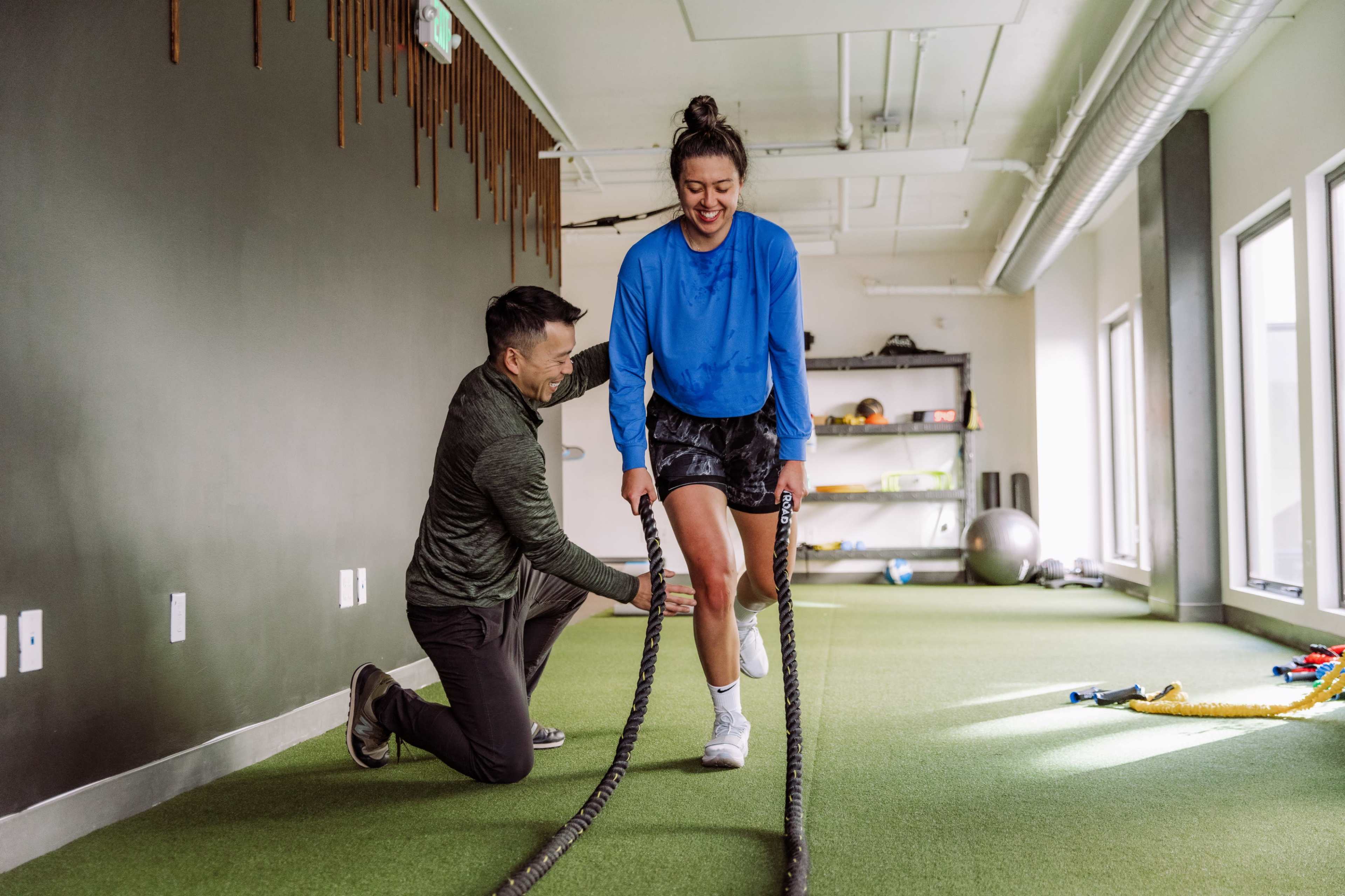 A trainer assists a woman in a workout using battle ropes in a gym setting.