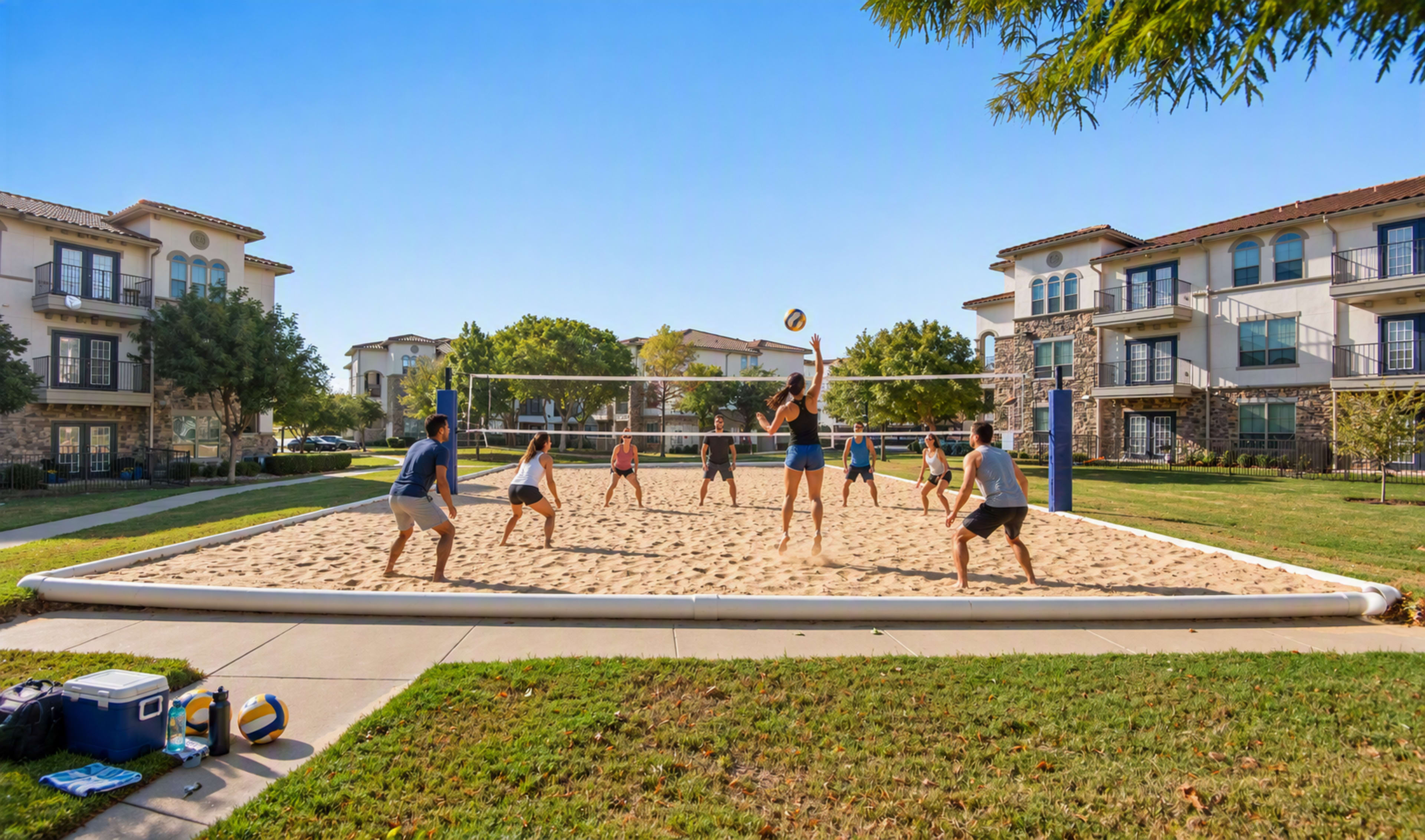 A group of people is playing beach volleyball on a sand court surrounded by residential buildings and green grass.