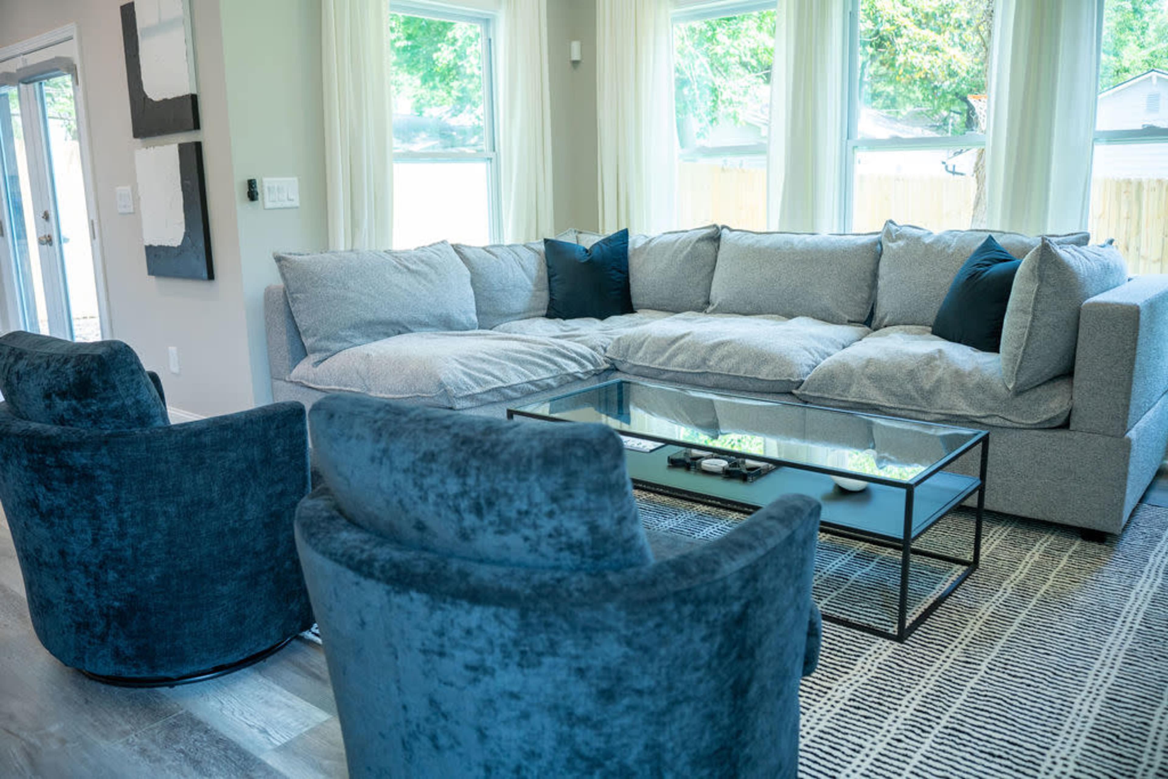 A modern living room with a large gray sectional sofa, two blue armchairs, and a glass coffee table placed on a patterned rug.