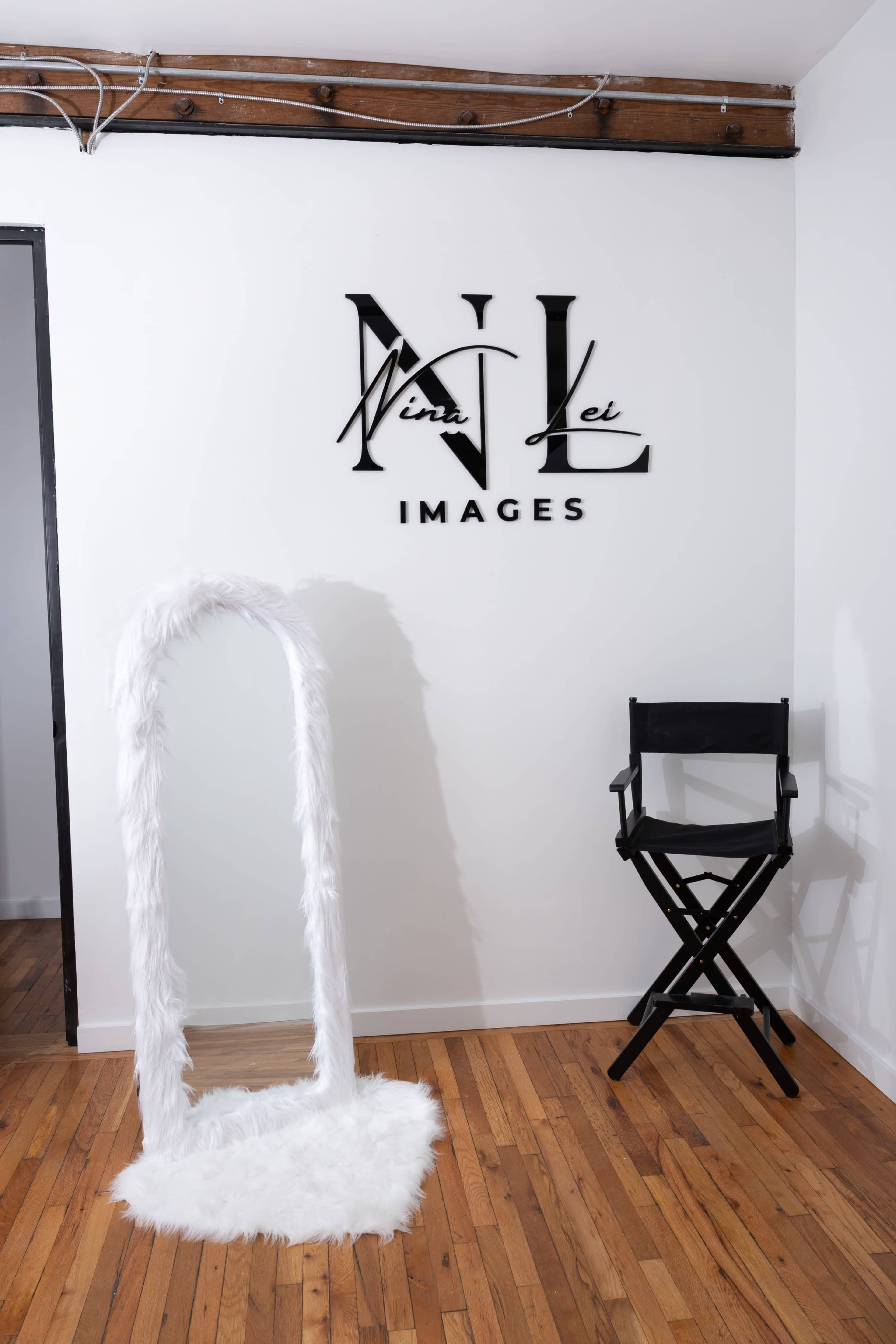 The image shows a minimalist studio interior featuring a white arch adorned with fluffy material and a black director's chair against a backdrop of a white wall with a decorative sign reading "N L IMAGES."