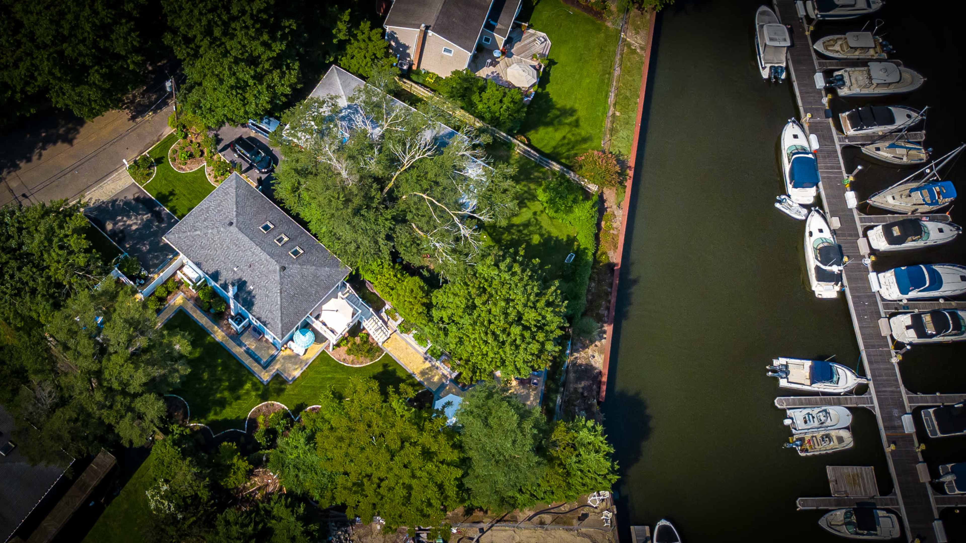 An aerial view shows a house surrounded by greenery with a marina and several boats docked nearby.