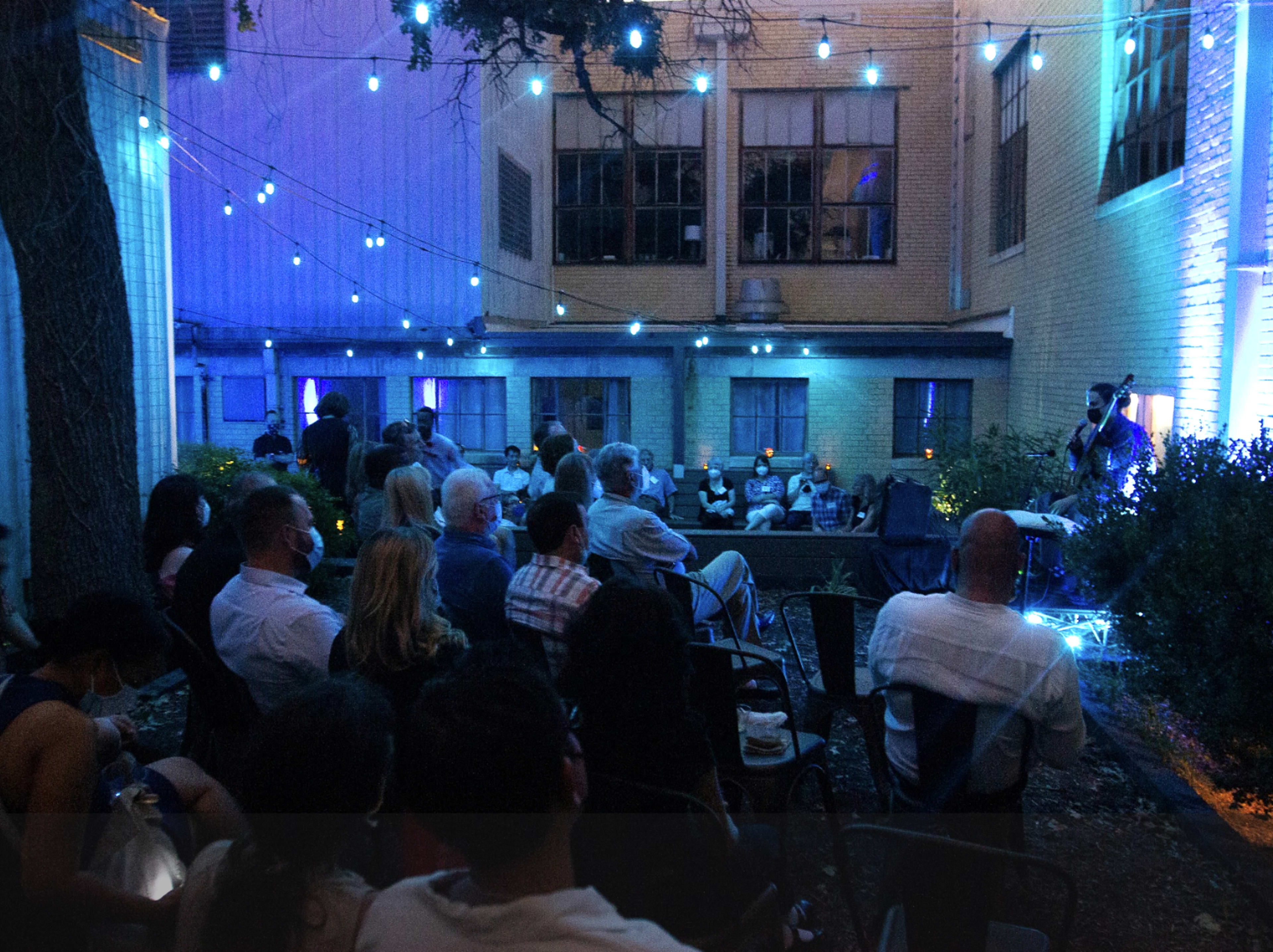 A crowd of people sits in an outdoor space illuminated by string lights, watching a performer on a stage.