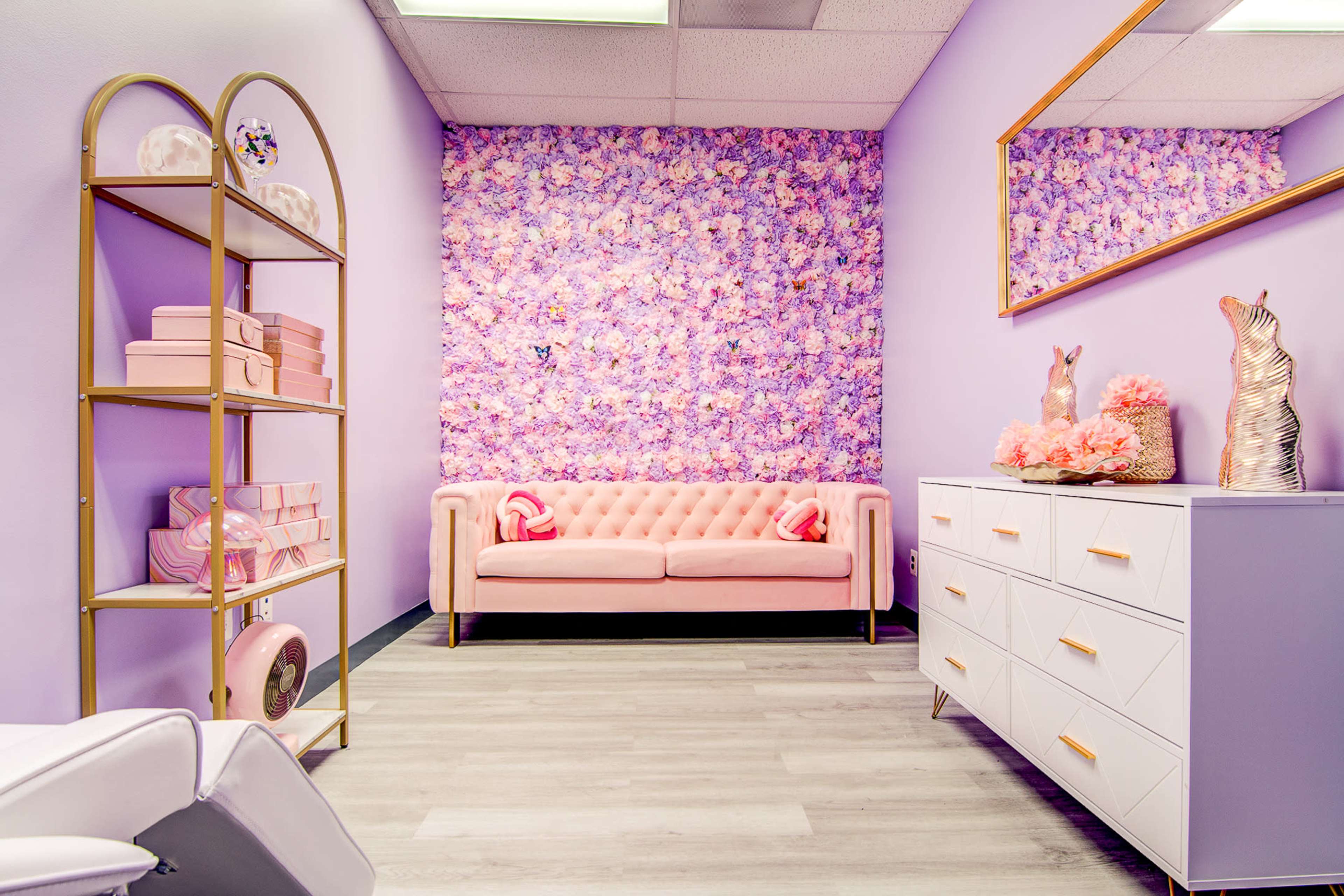 A pastel-themed room with a pink tufted sofa, a floral wall, and decorative furnishings including a gold shelving unit and a white dresser.