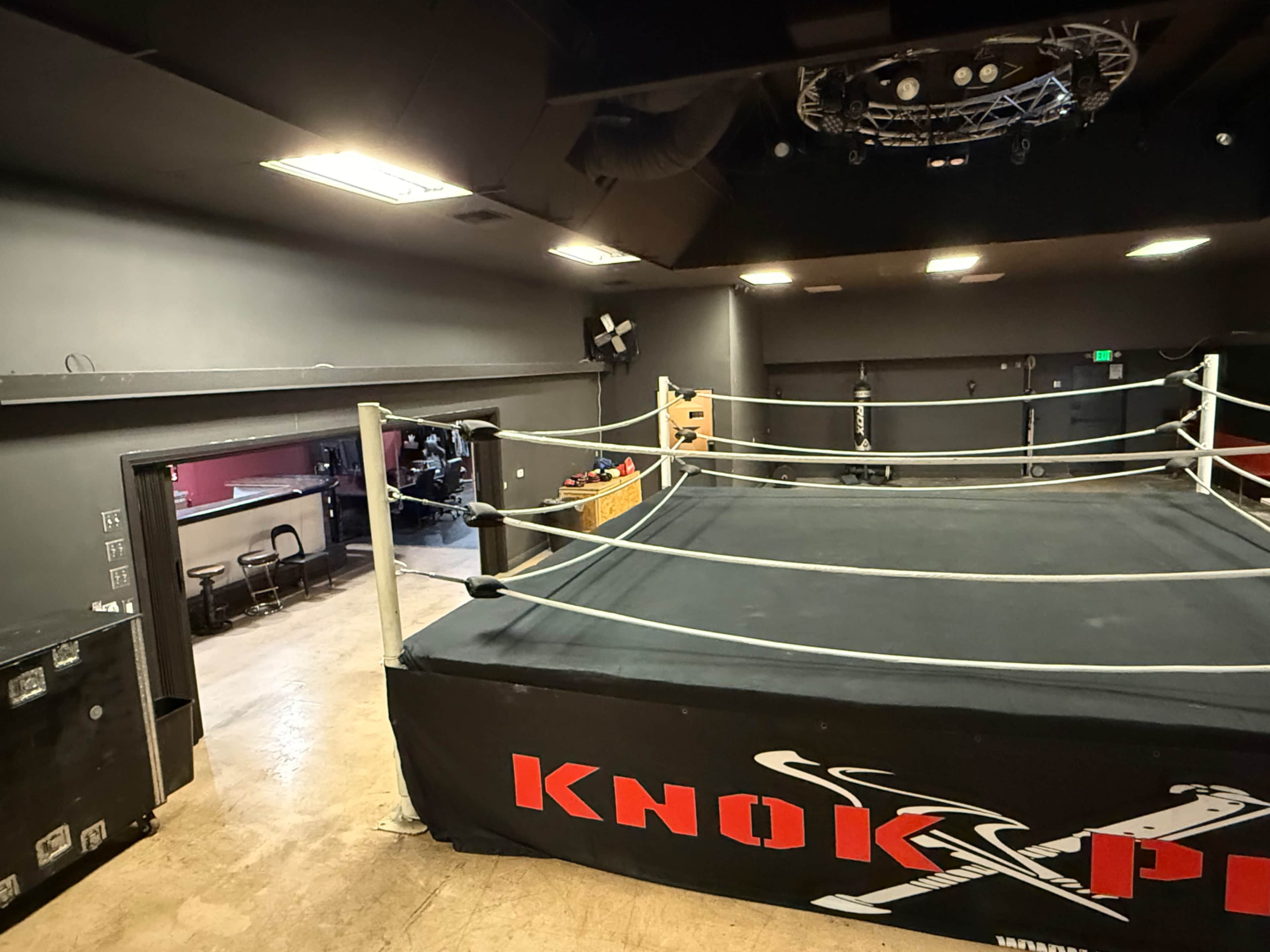The image shows a wrestling ring in a dimly lit room with a gray wall and some equipment visible in the background.