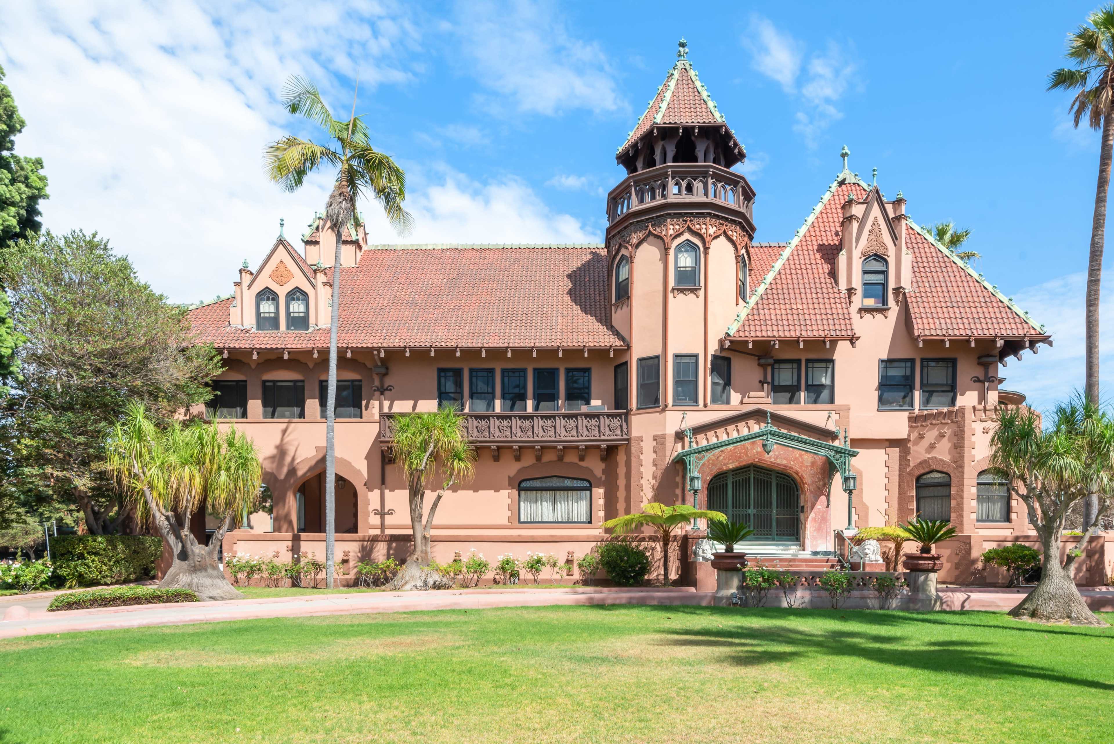 The image shows a large, ornate pink mansion with a turret, intricate architectural details, and a well-maintained lawn surrounded by palm trees.