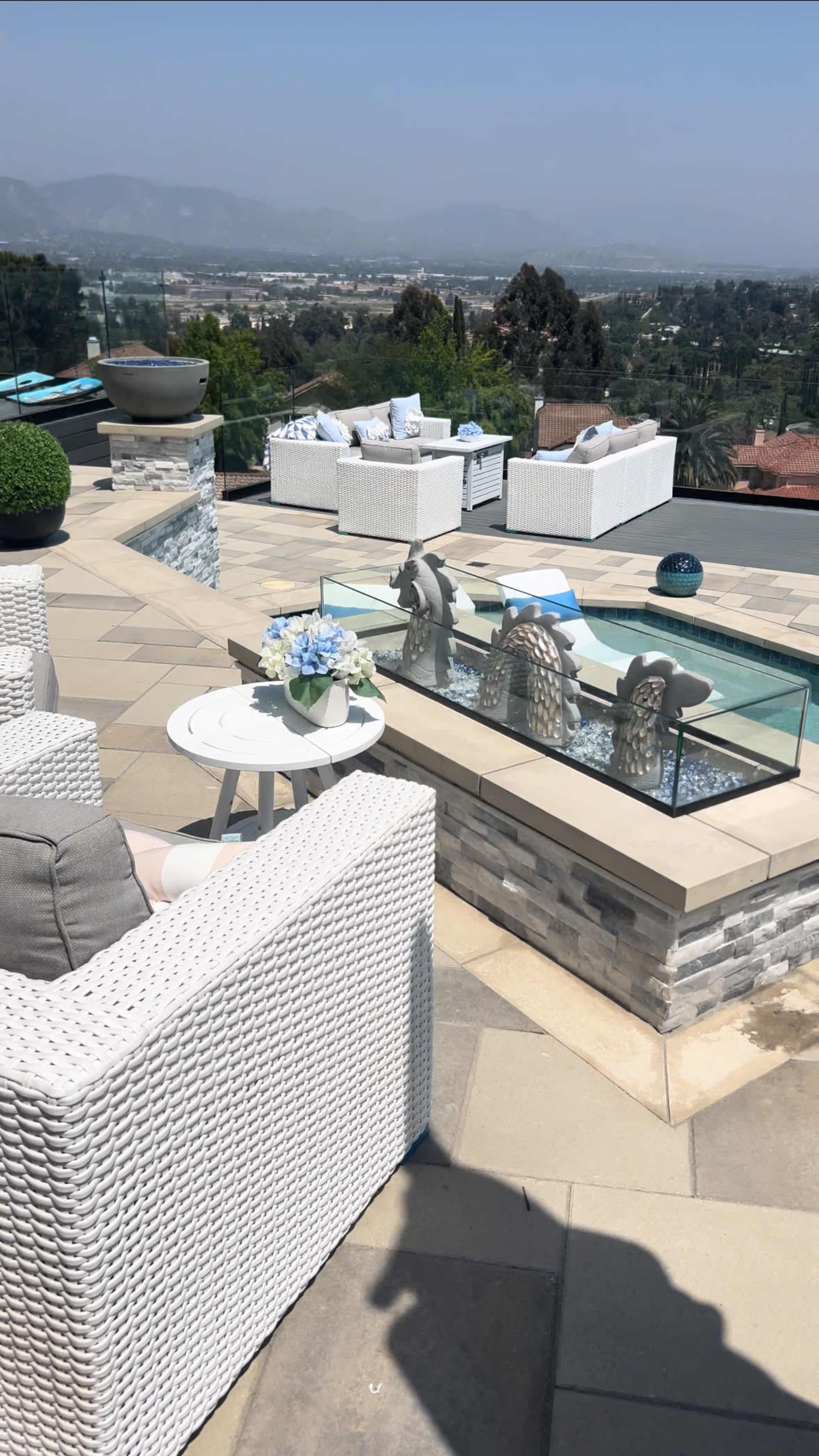 A modern outdoor patio featuring white wicker furniture, a glass-topped table with flowers, and a small water feature with decorative elements.