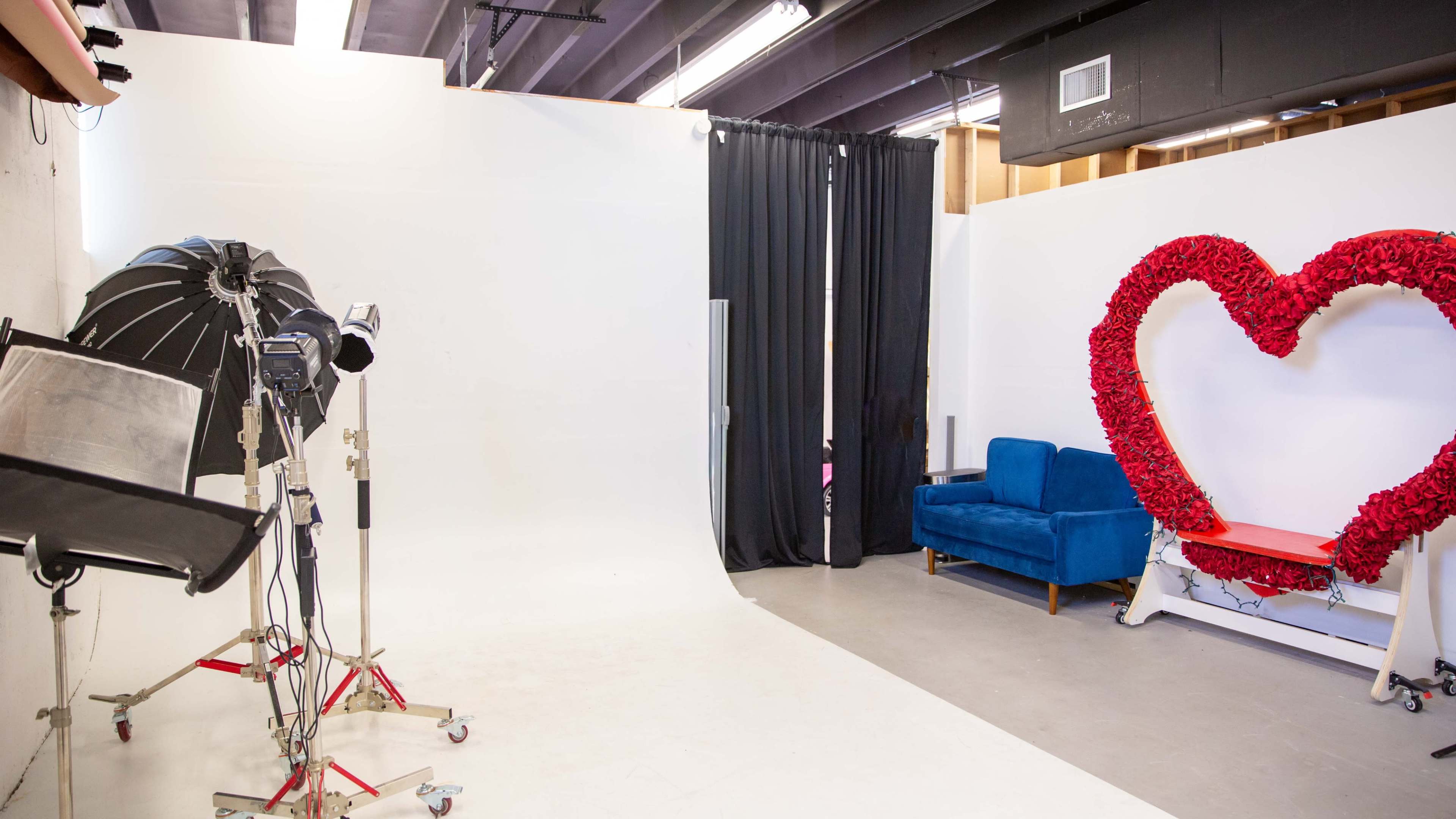 The image shows a spacious photography studio with a white backdrop, a blue couch, and a large heart-shaped decoration made of red flowers.