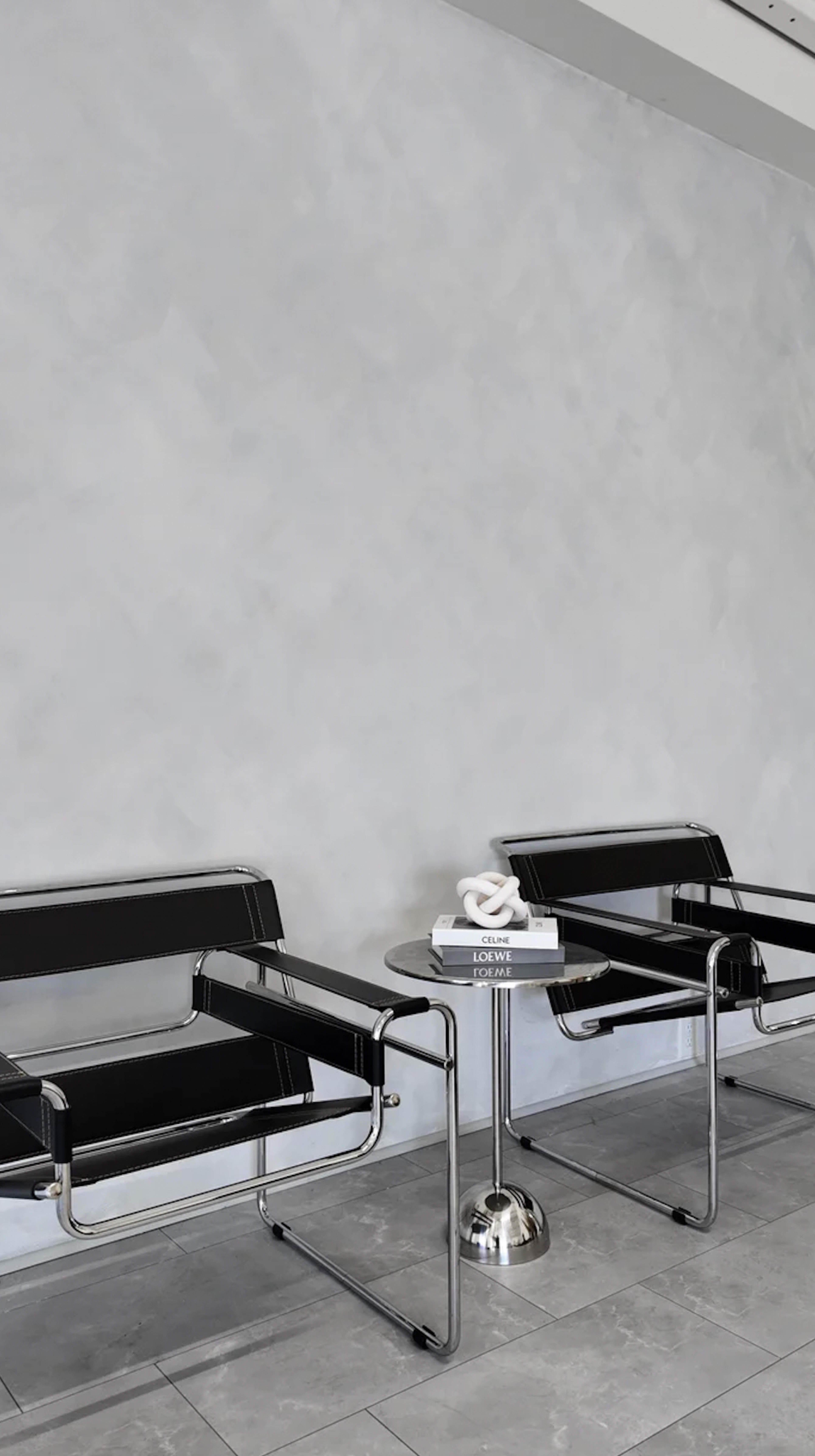 The image shows two modern black chairs with chrome frames positioned near a small round table that holds a stack of books and a decorative object.