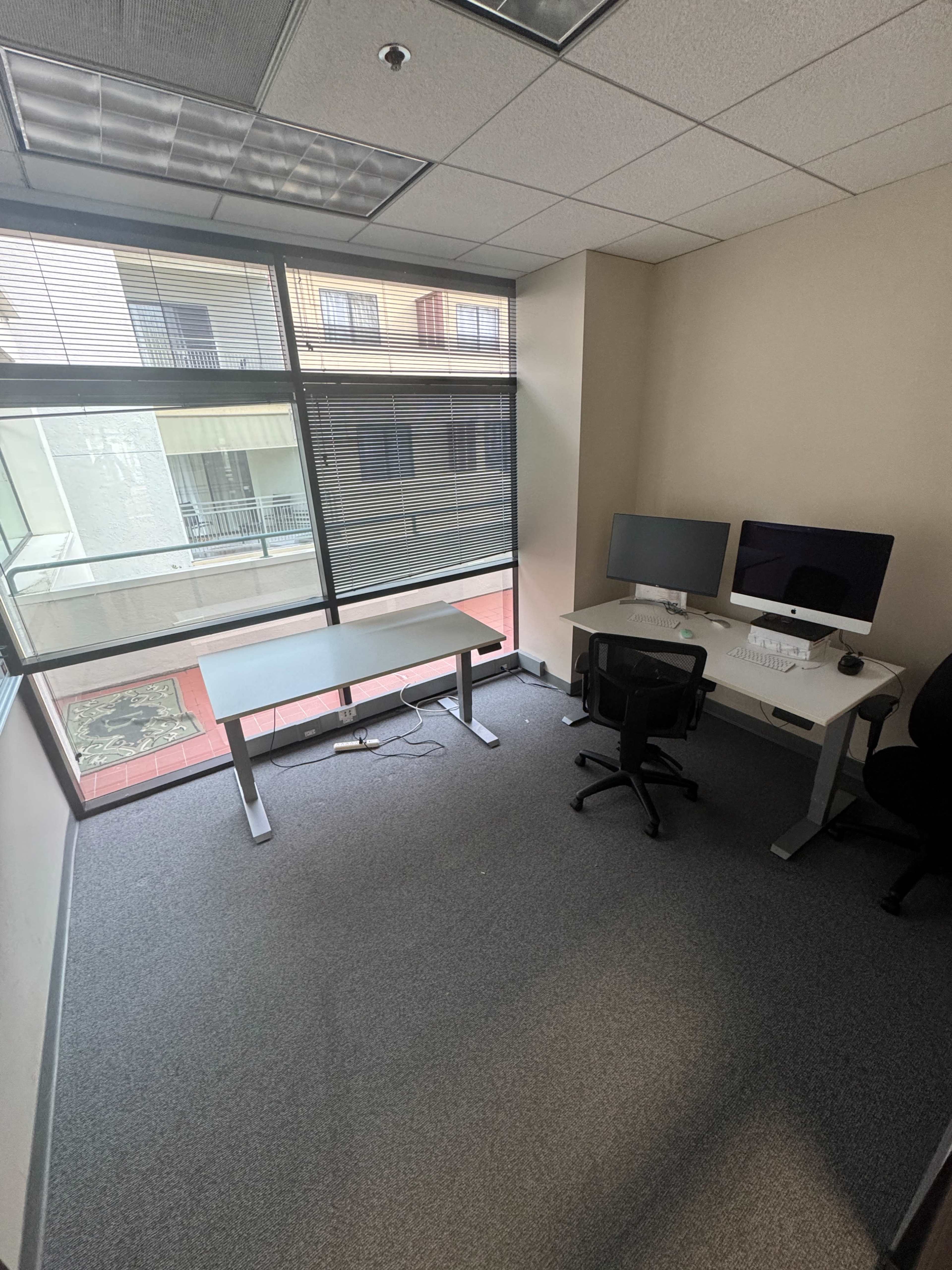 The image shows a sparse office space with two computer monitors, a desk, a rolling chair, and large windows allowing natural light.