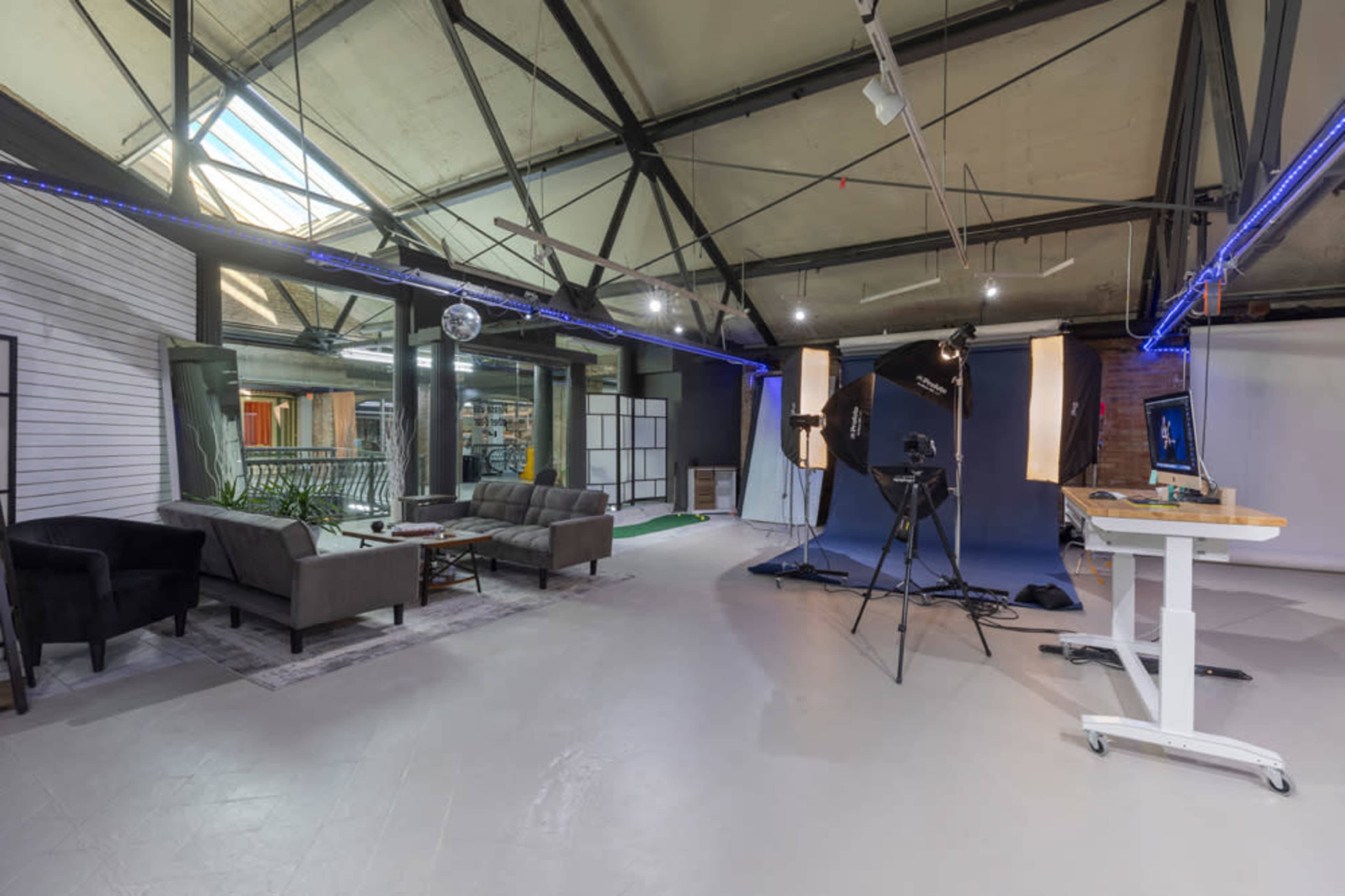A spacious studio with a modern layout featuring a lounge area with sofas, a photography setup with lighting equipment, and large windows allowing natural light to enter.