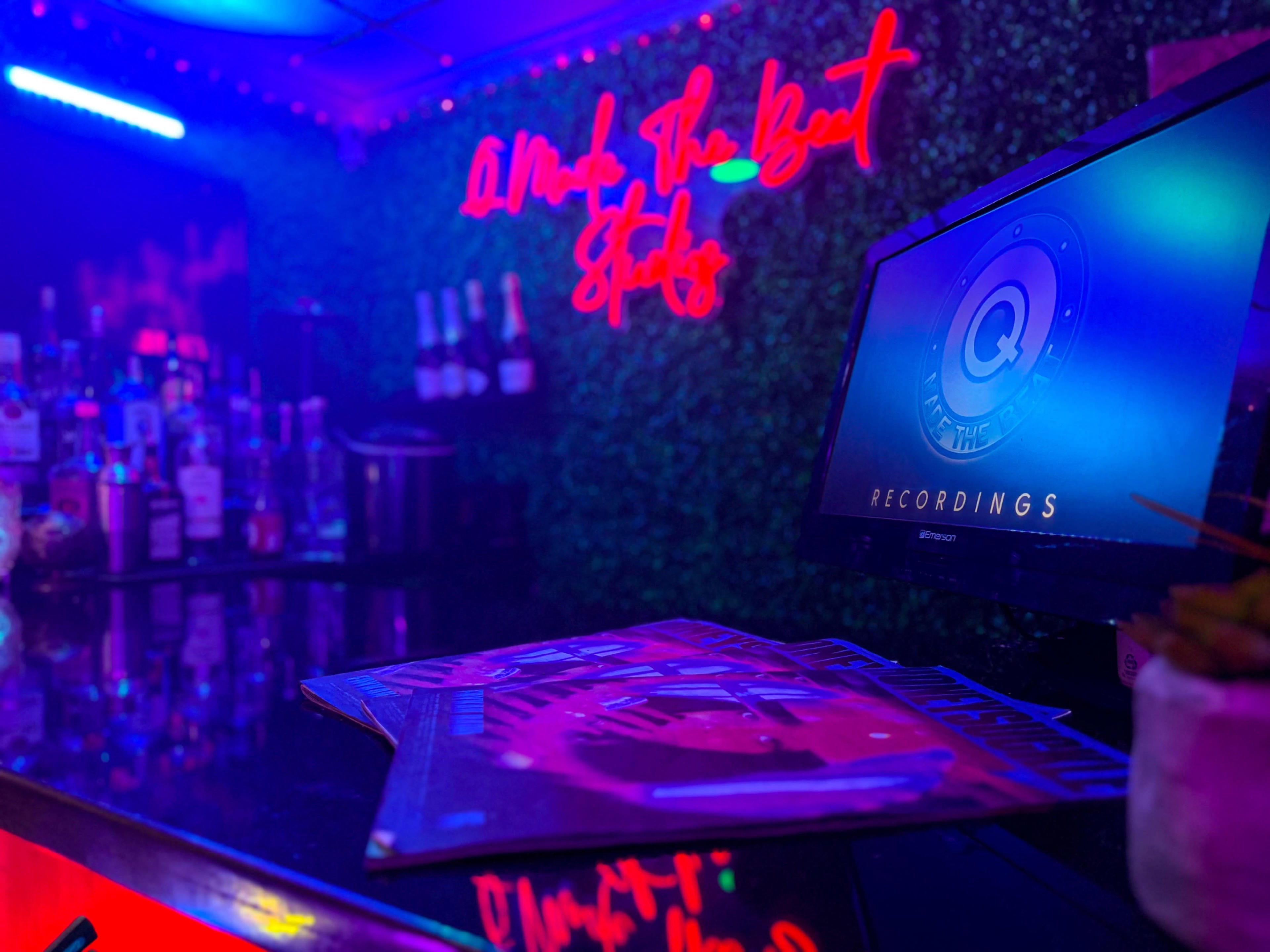 A dimly lit bar display features a monitor showing "Q The Beat Recordings" against a backdrop of illuminated bottles and neon signs.