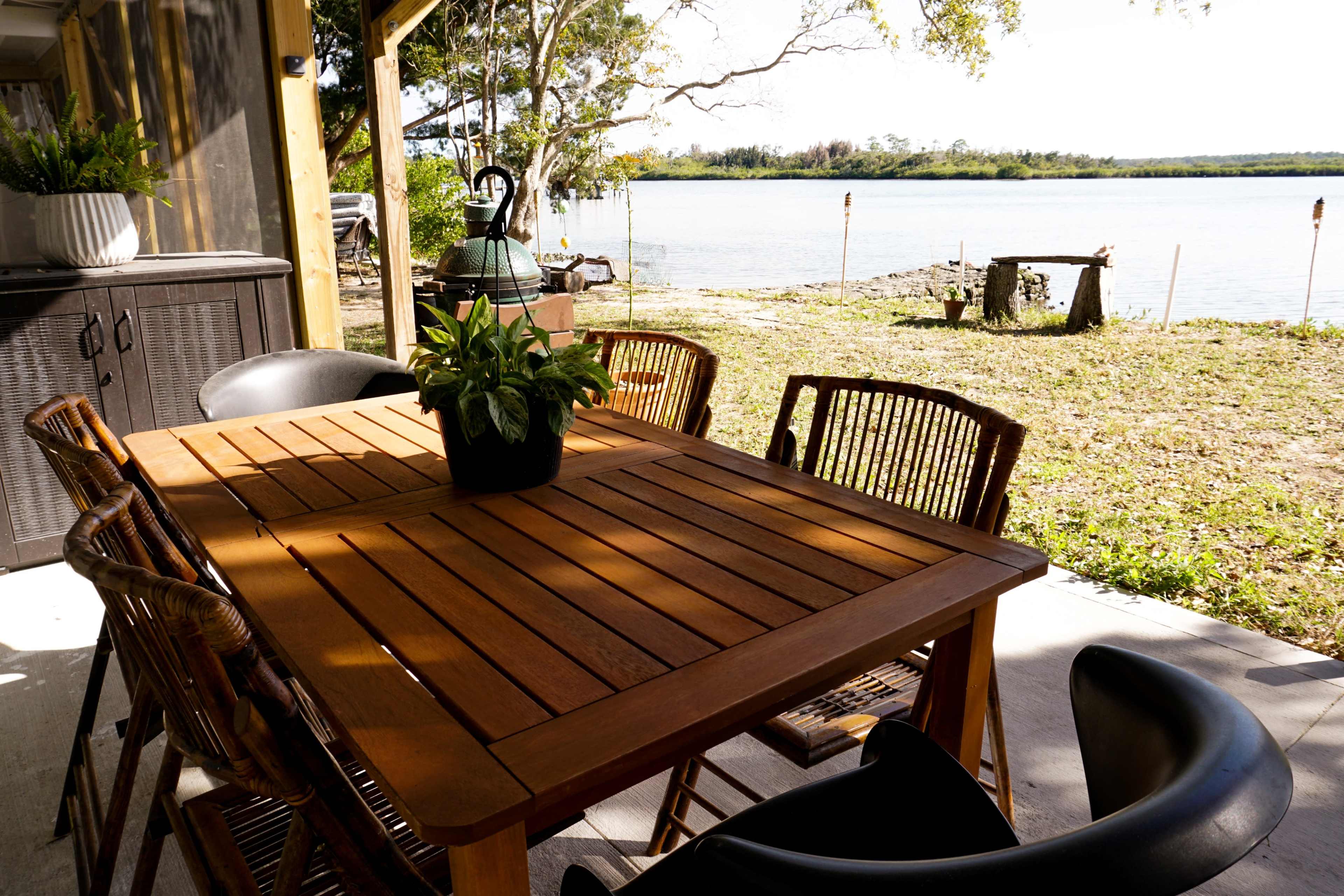 A wooden dining table with chairs is set outdoors near a riverbank, surrounded by greenery.