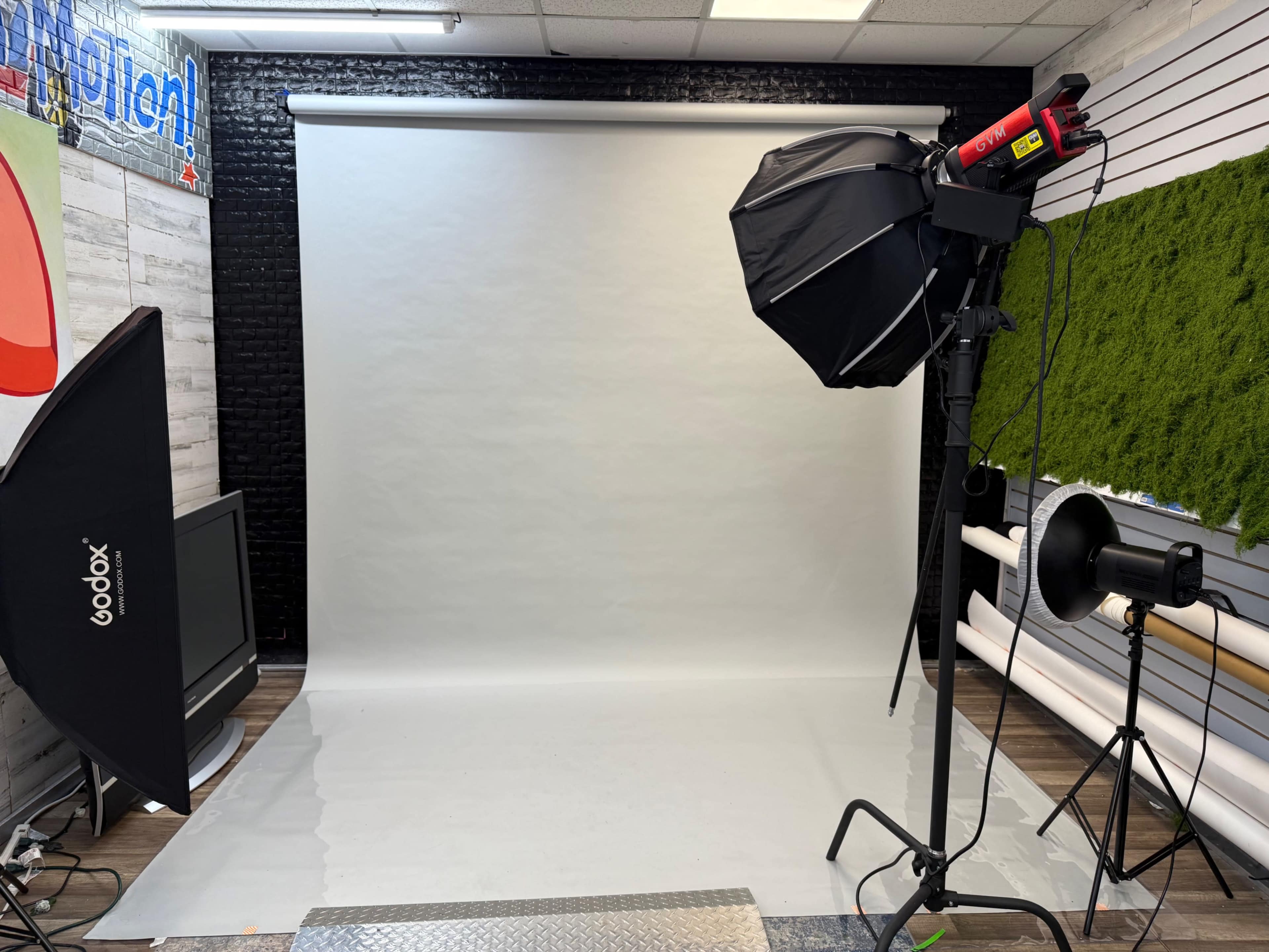 The image shows a photography studio with a plain backdrop, lighting equipment, and a television set.