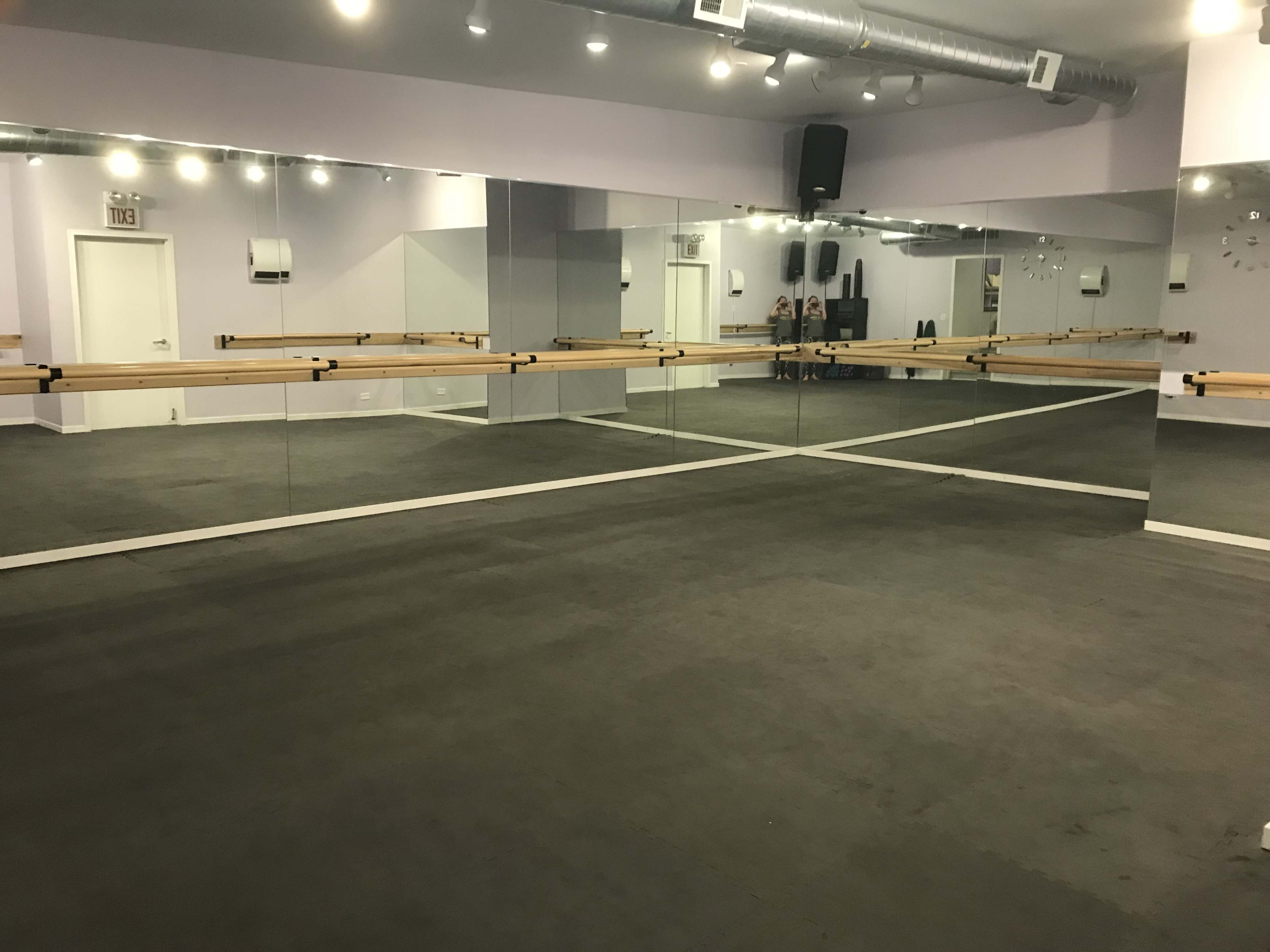 A dance studio featuring mirrored walls, a wooden ballet barre, and a grey floor.