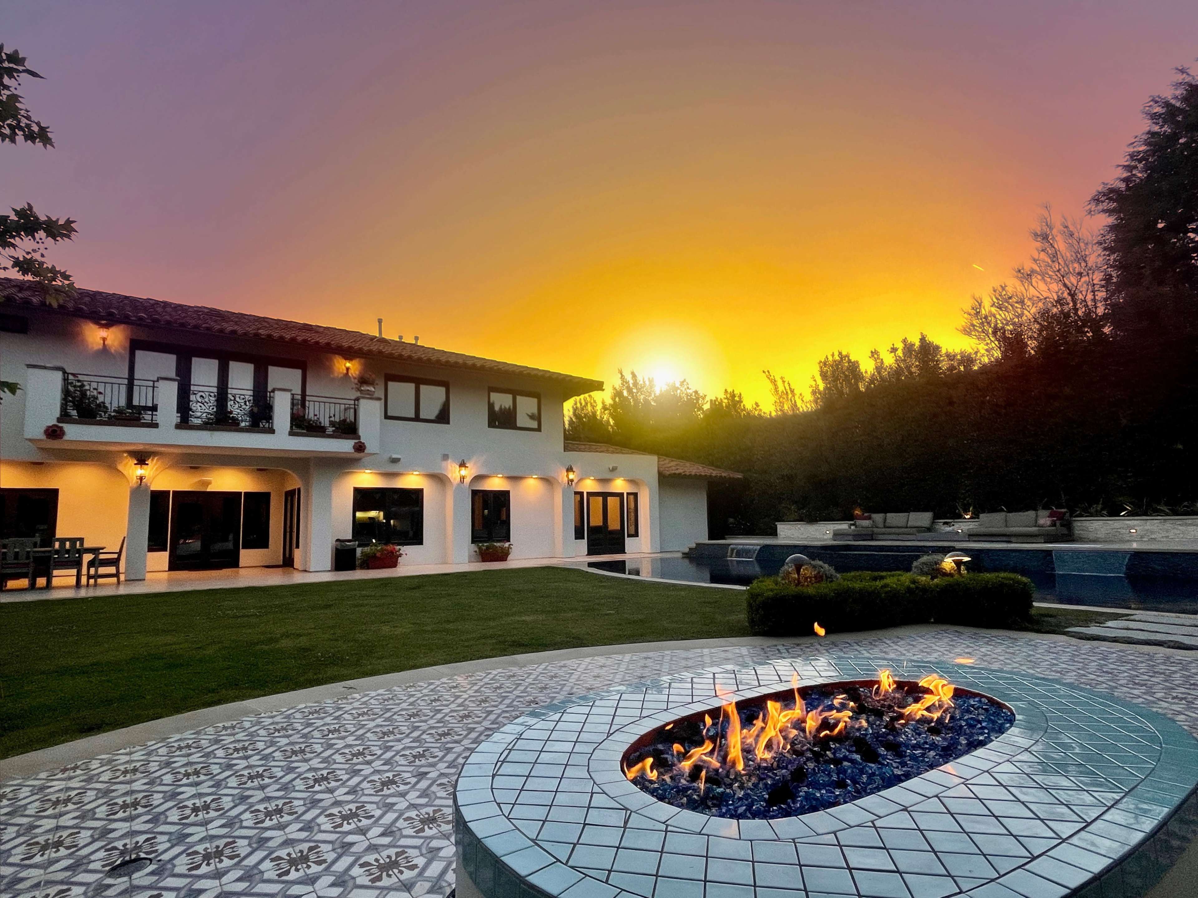 A backyard features a gas fire pit surrounded by decorative tiles, with a view of a modern house and a sunset casting an orange light.