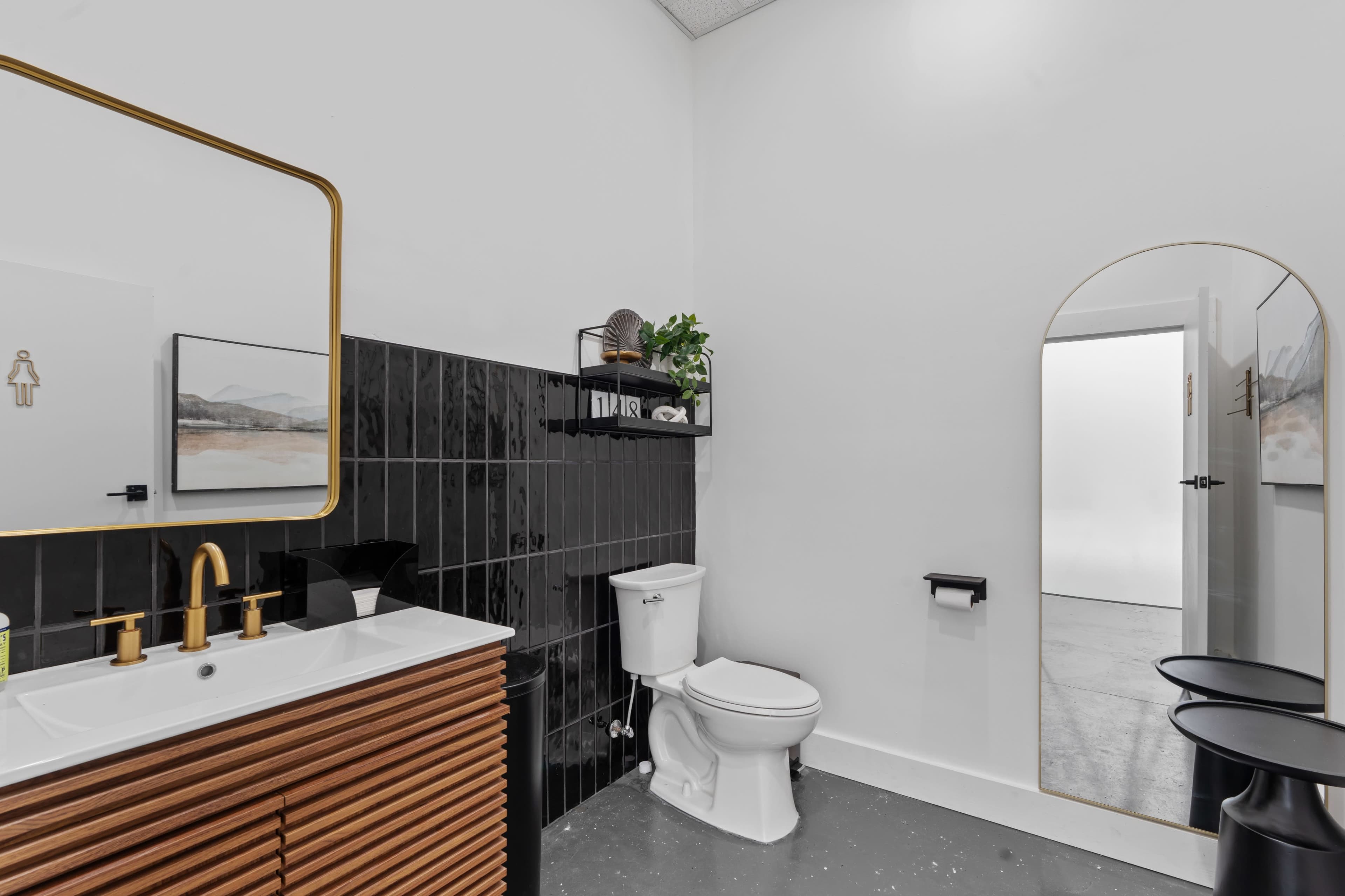 The bathroom features a wooden vanity with a sink, a wall-mounted mirror with a gold frame, a toilet, and a small shelf with decor, set against black tiled walls.