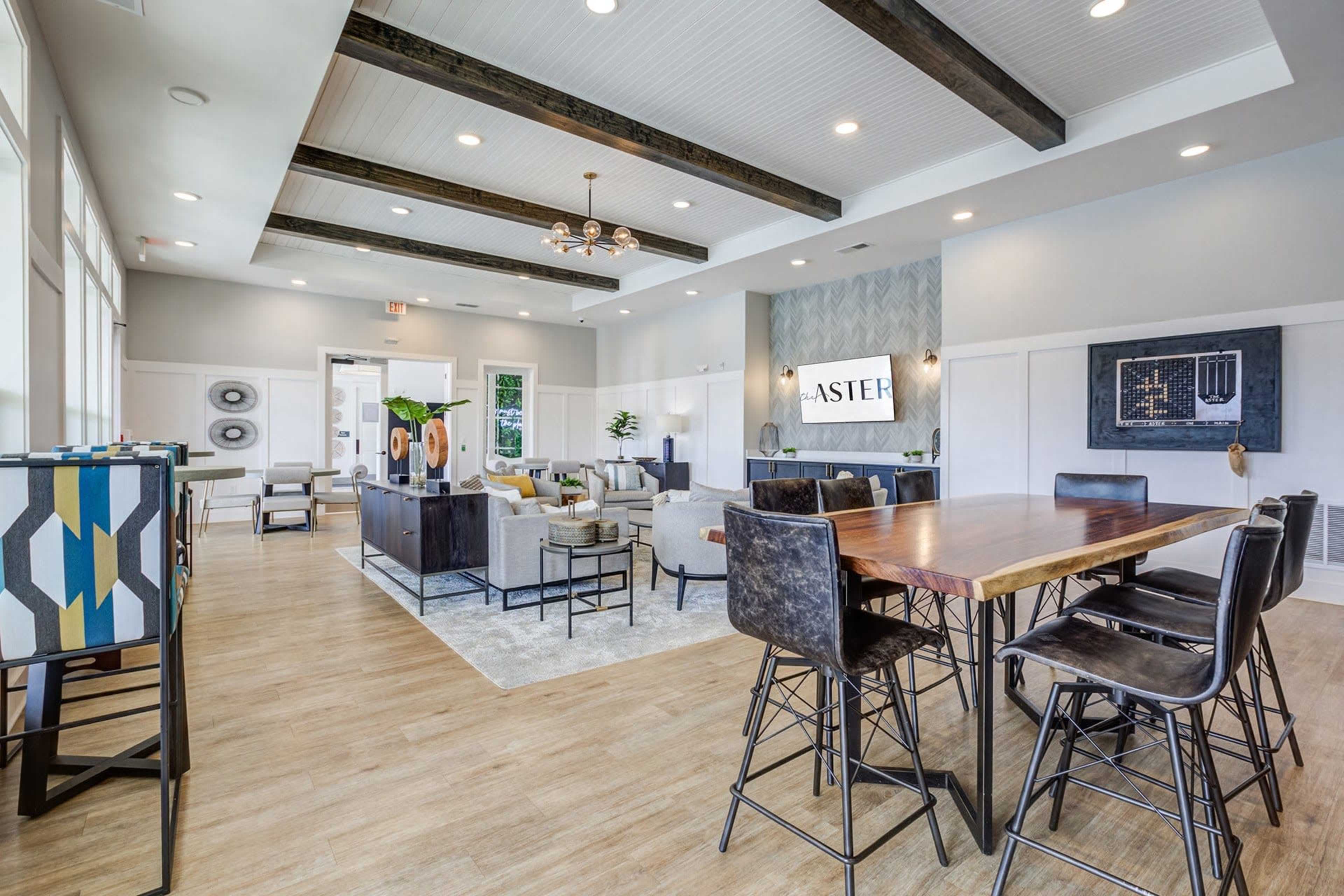 An interior lounge area with modern furniture, wooden beams on the ceiling, and a large communal table surrounded by high chairs.