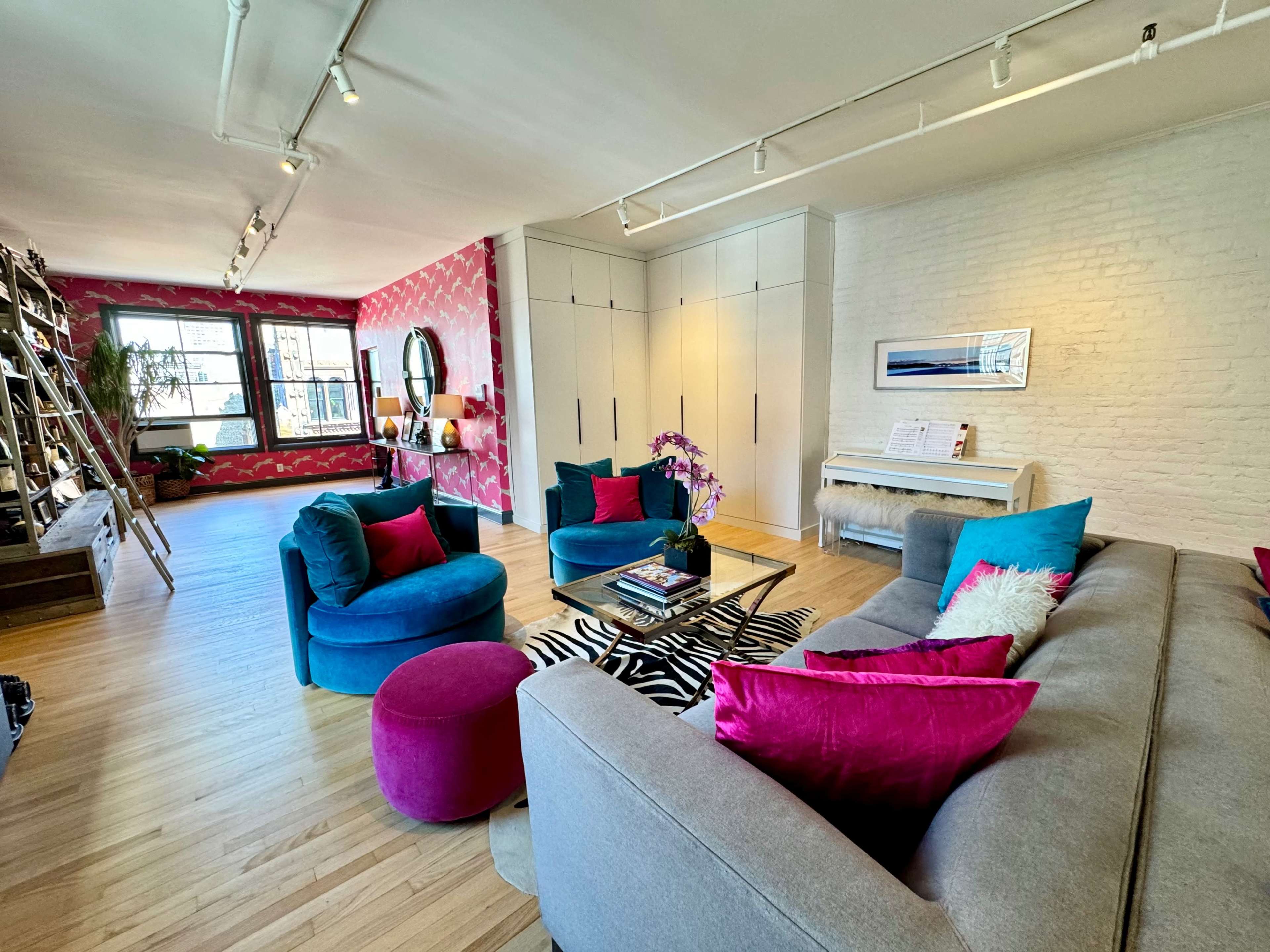The image shows a modern living room with a gray sofa, colorful accent pillows, a round coffee table, a piano, and a decorative rug on a wooden floor.