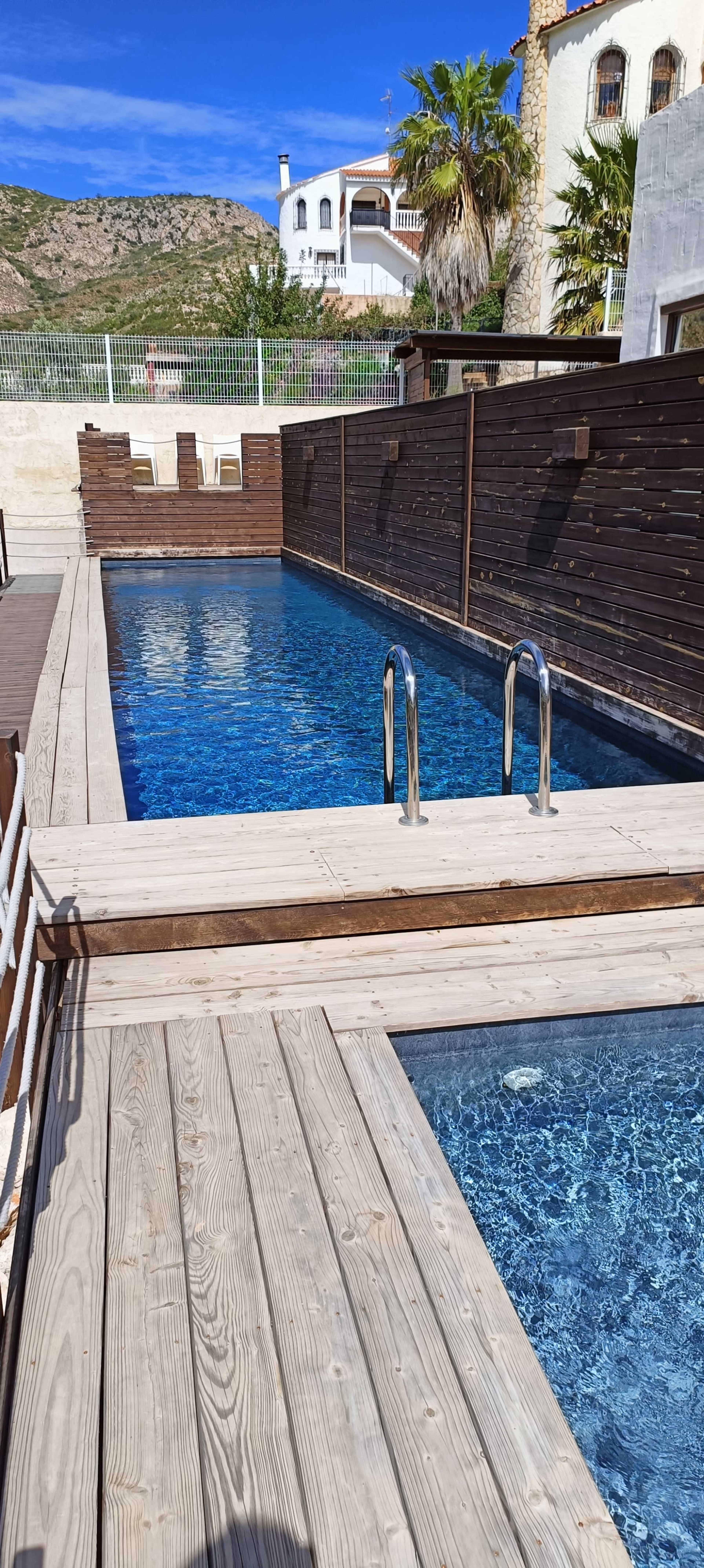 A clear swimming pool surrounded by wooden decking and a fence, with mountains visible in the background.