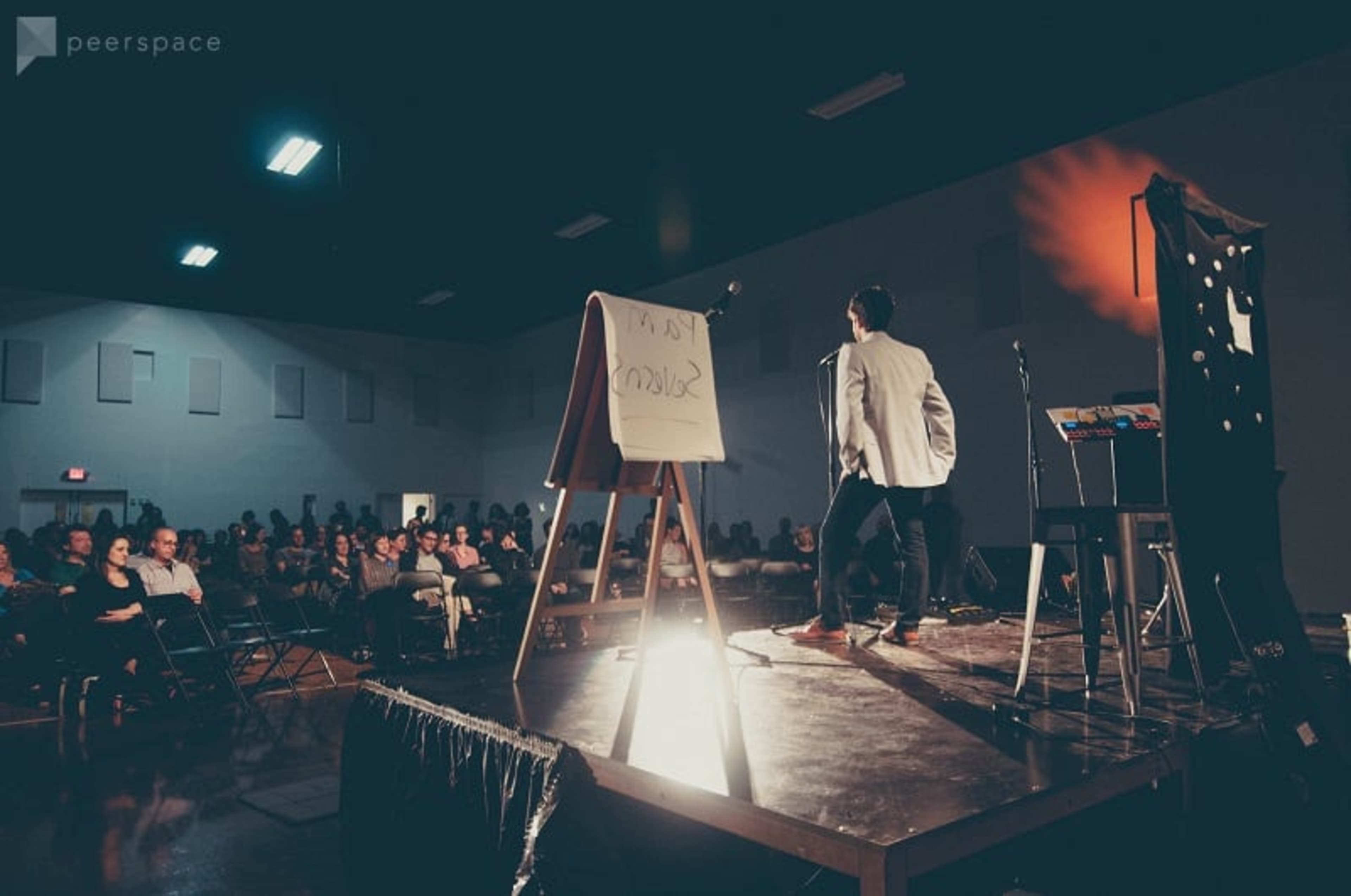 A speaker stands on stage in front of an audience, with a blank presentation board and equipment nearby.