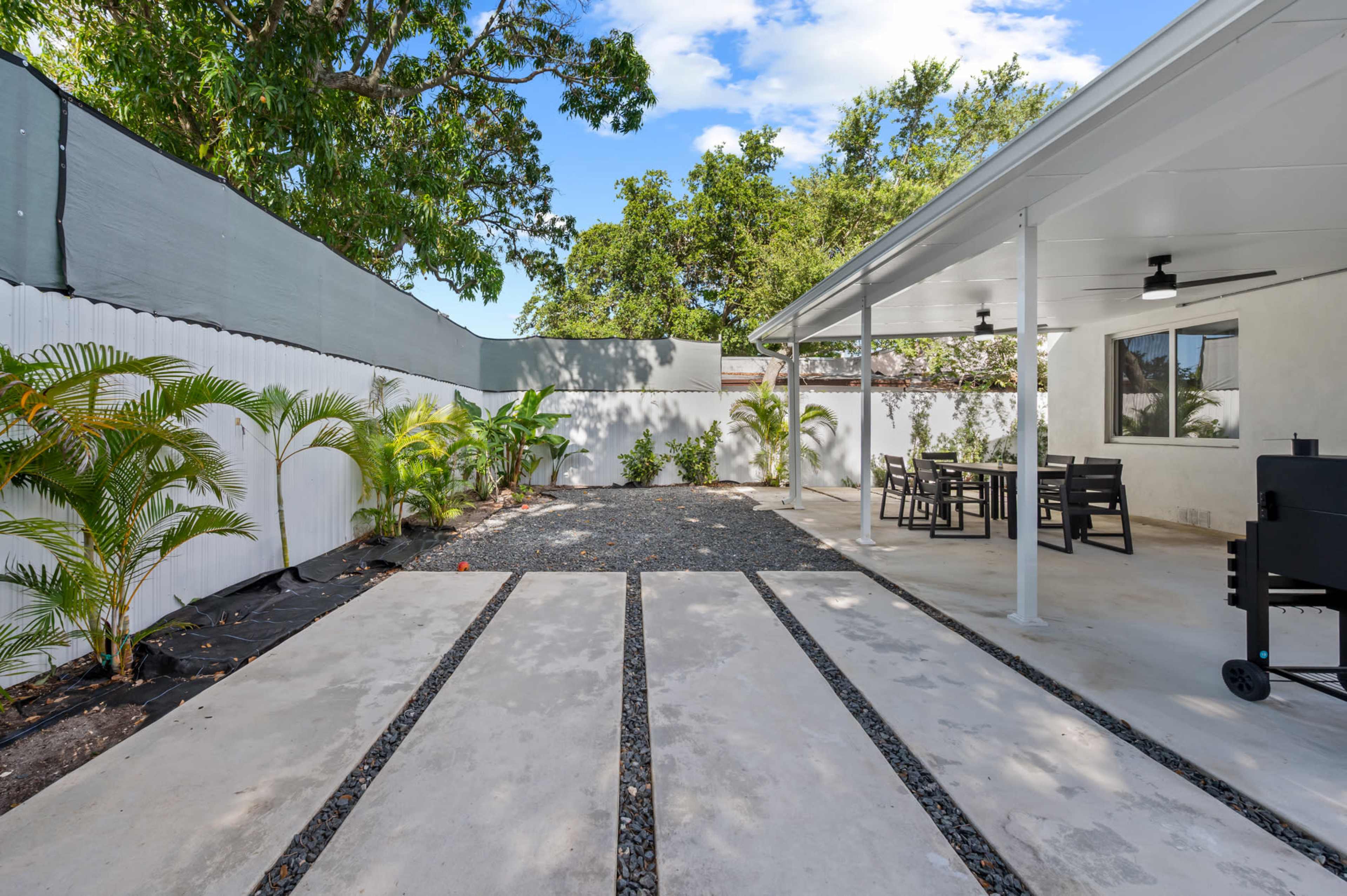 The image shows a patio area with gravel pathways, a dining table, and a grill, surrounded by greenery and a white fence.