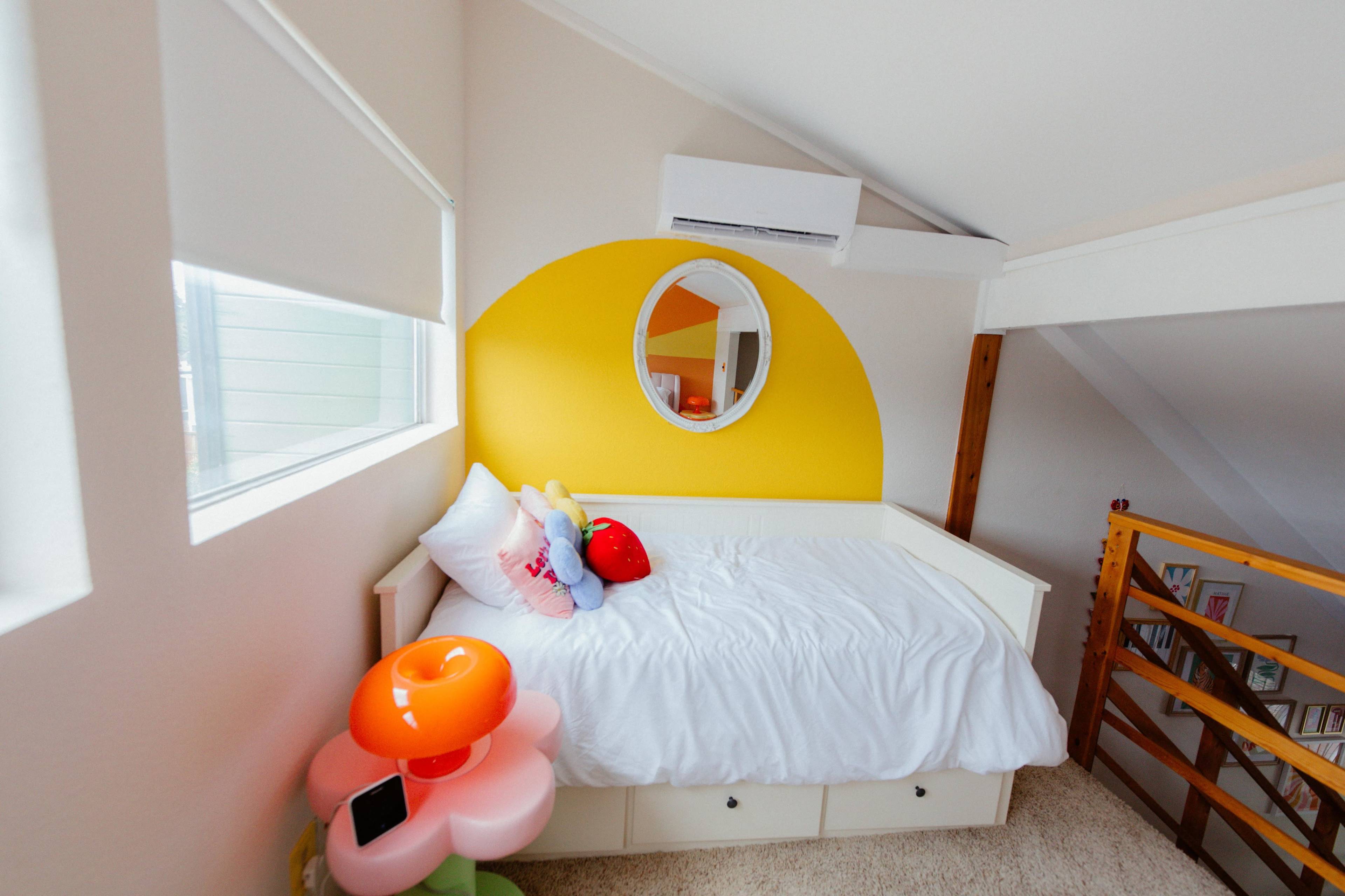 The image shows a cozy bedroom with a white bed against a bright yellow half-circle wall and colorful decorations, including a flower-shaped lamp.