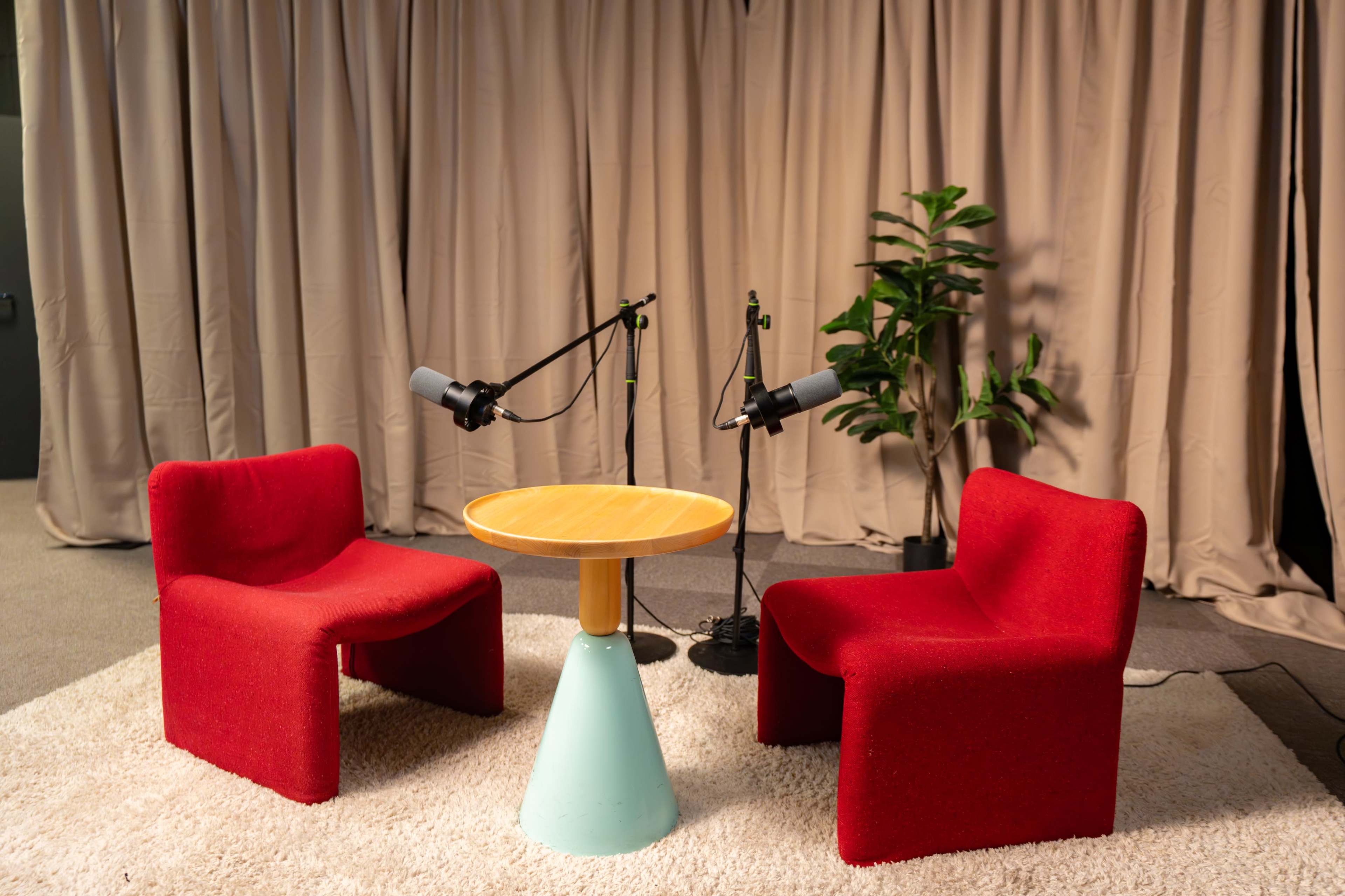 The image shows a cozy podcasting setup with two red chairs, a round wooden table, and two microphones on stands, all arranged in front of a beige curtain and a potted plant.
