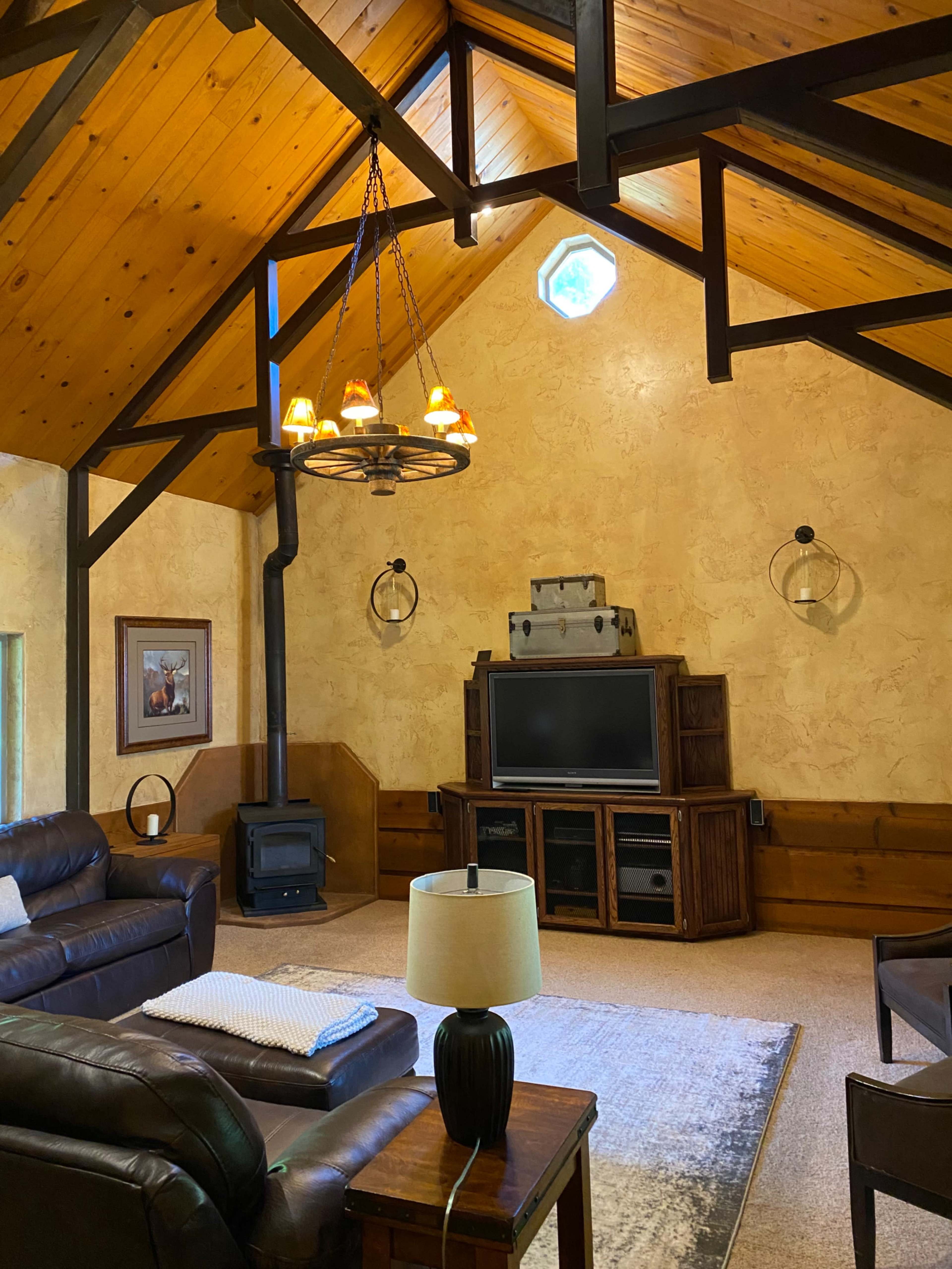 A spacious living room features a high wooden ceiling, a wall-mounted television, a cozy seating area, and a wood-burning stove.