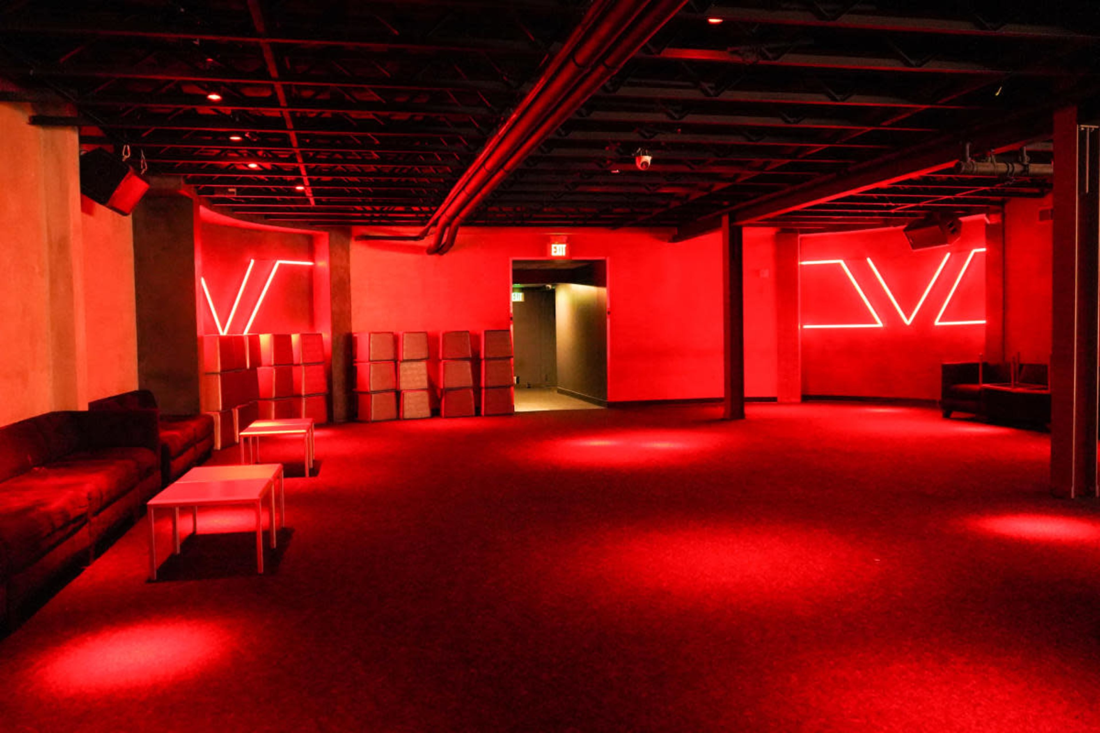 The image shows a dimly lit interior space with red lighting, featuring a few couches and tables, and a simplistic design with a minimalist style.