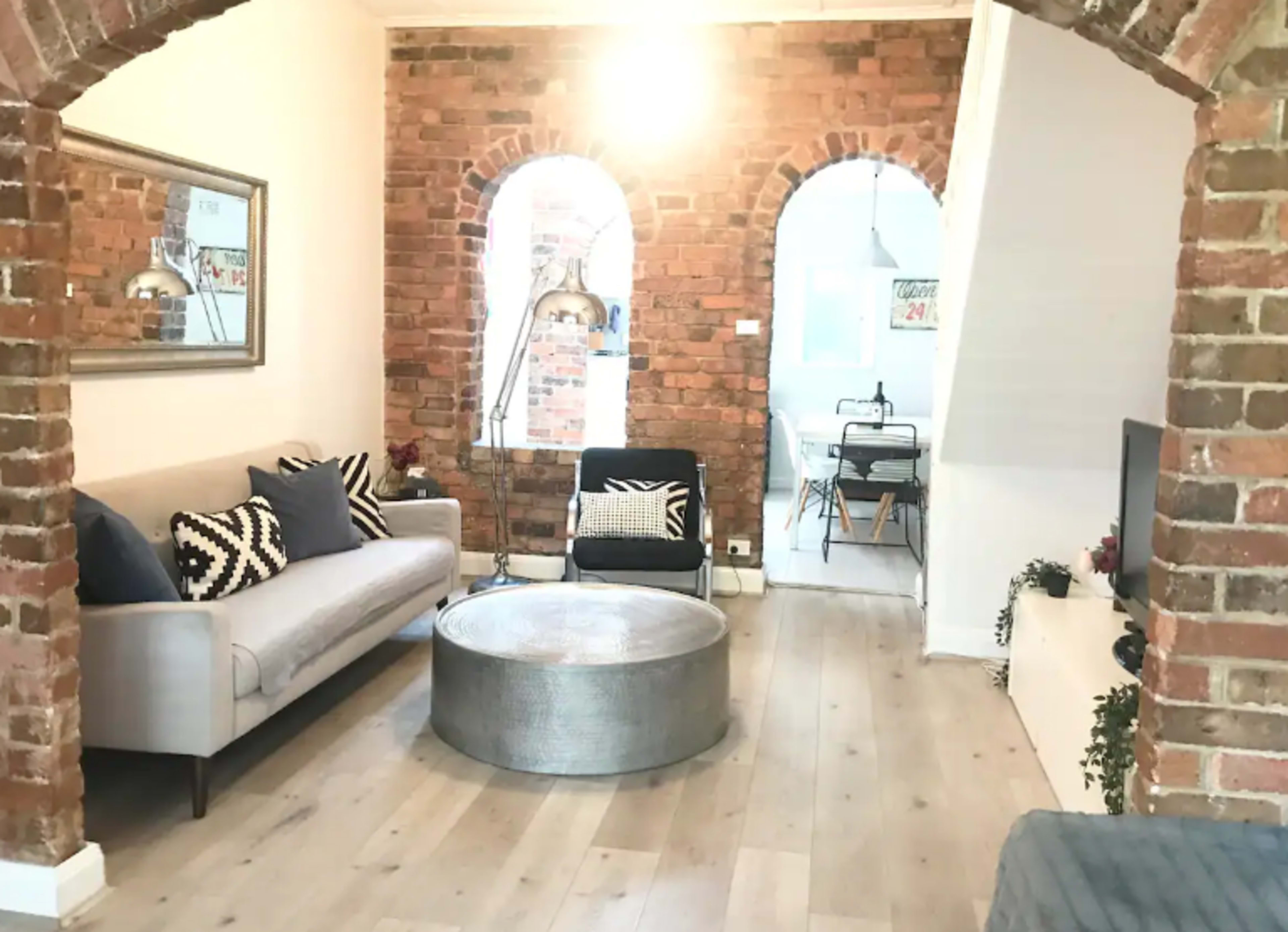 A living room with exposed brick walls, a gray sofa, and a round metallic coffee table, leading into a dining area visible in the background.