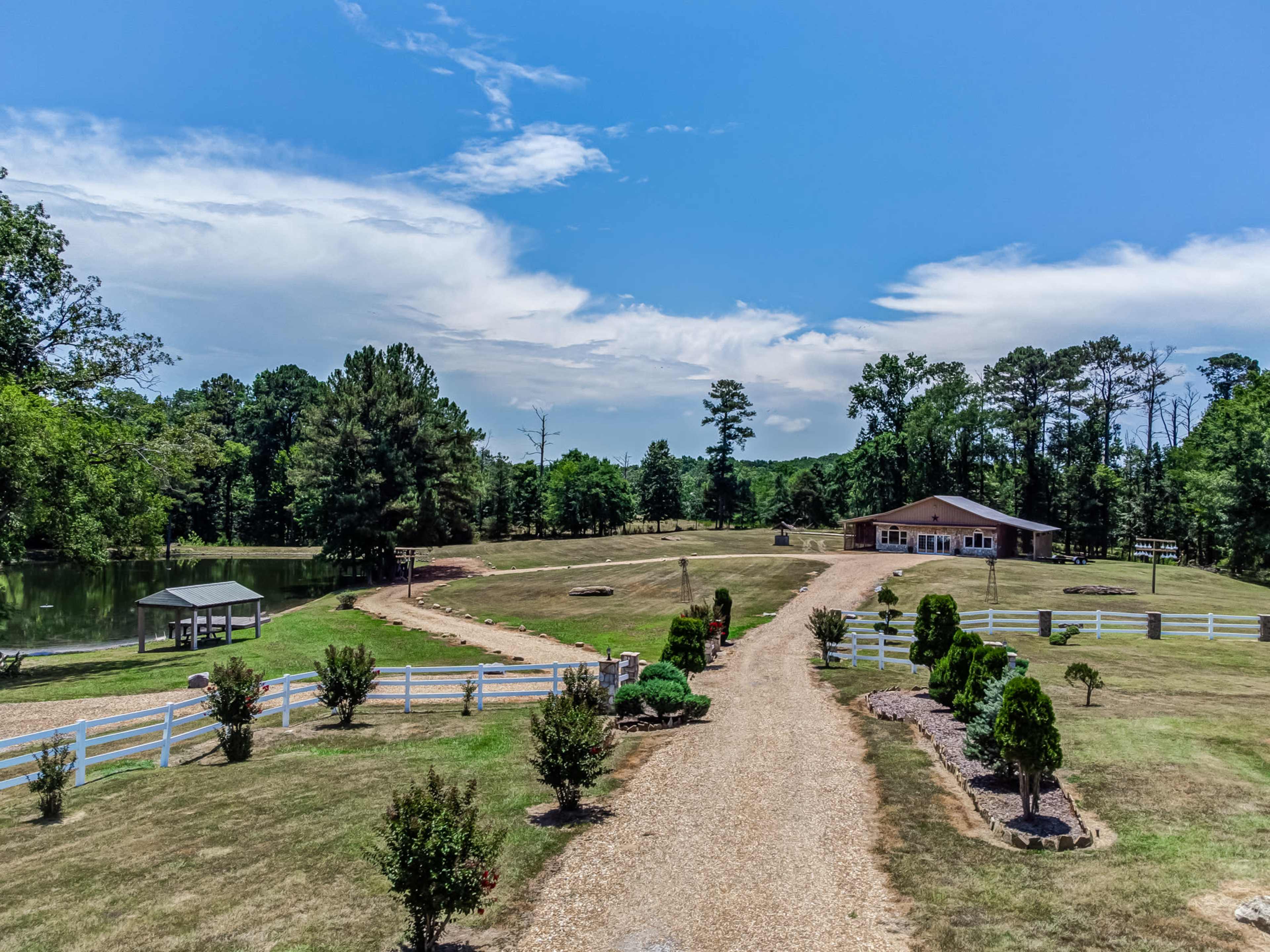 The image shows a spacious landscape with a gravel driveway leading to a large building, surrounded by manicured greenery and a pond on the left.