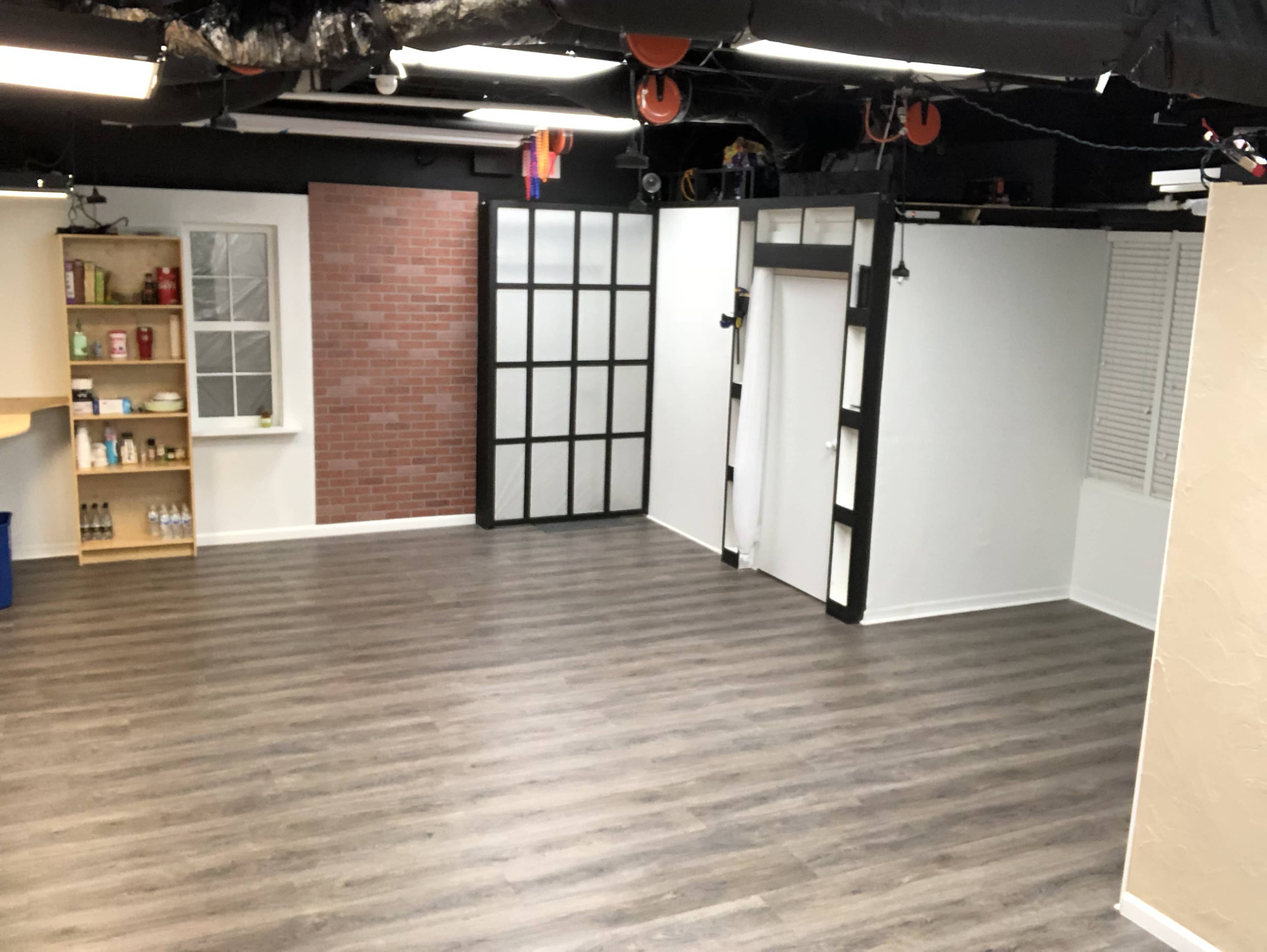 An empty room with a wooden floor, featuring exposed walls with a brick section, glass-paneled doors, and a shelf stocked with items against one wall.
