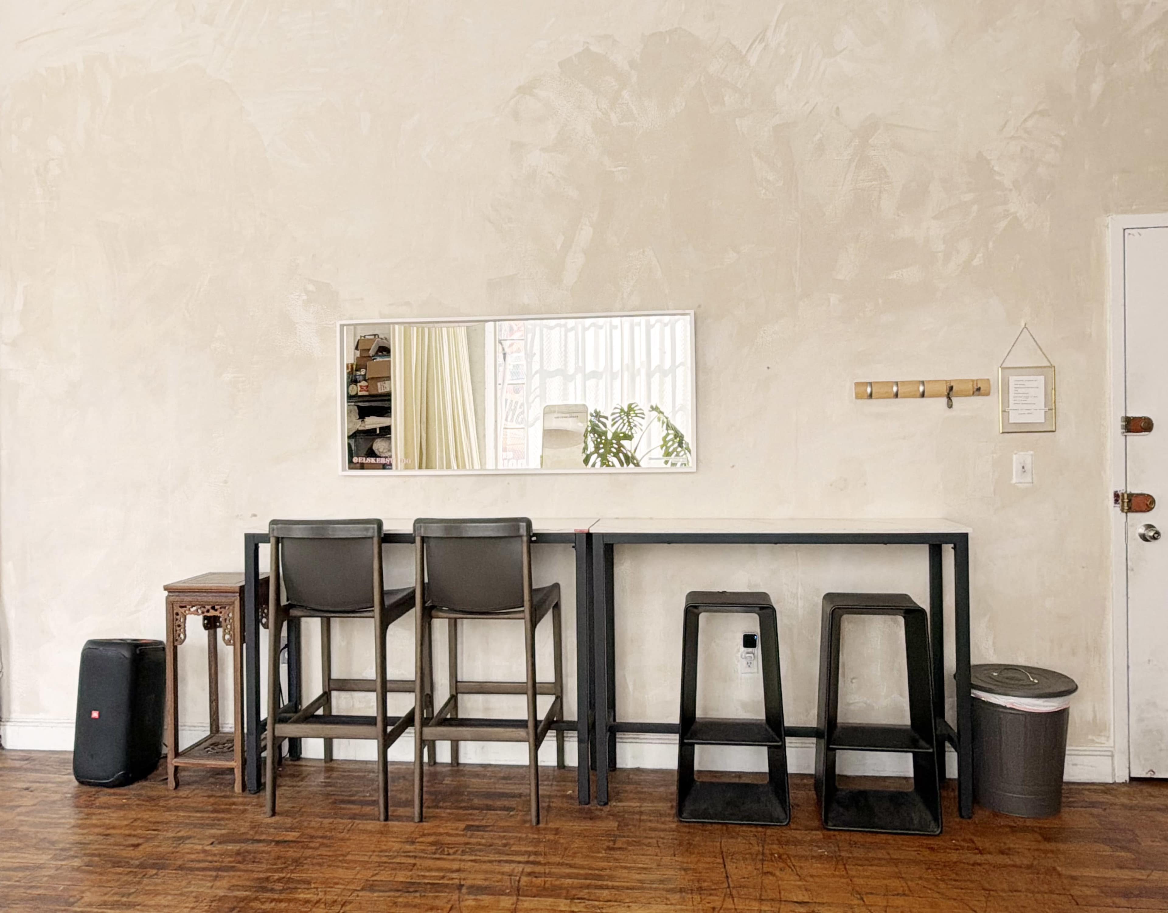 The image shows a minimalist interior with a wall-mounted mirror above a long table paired with three high stools, two stackable stools nearby, and a trash can in a corner.
