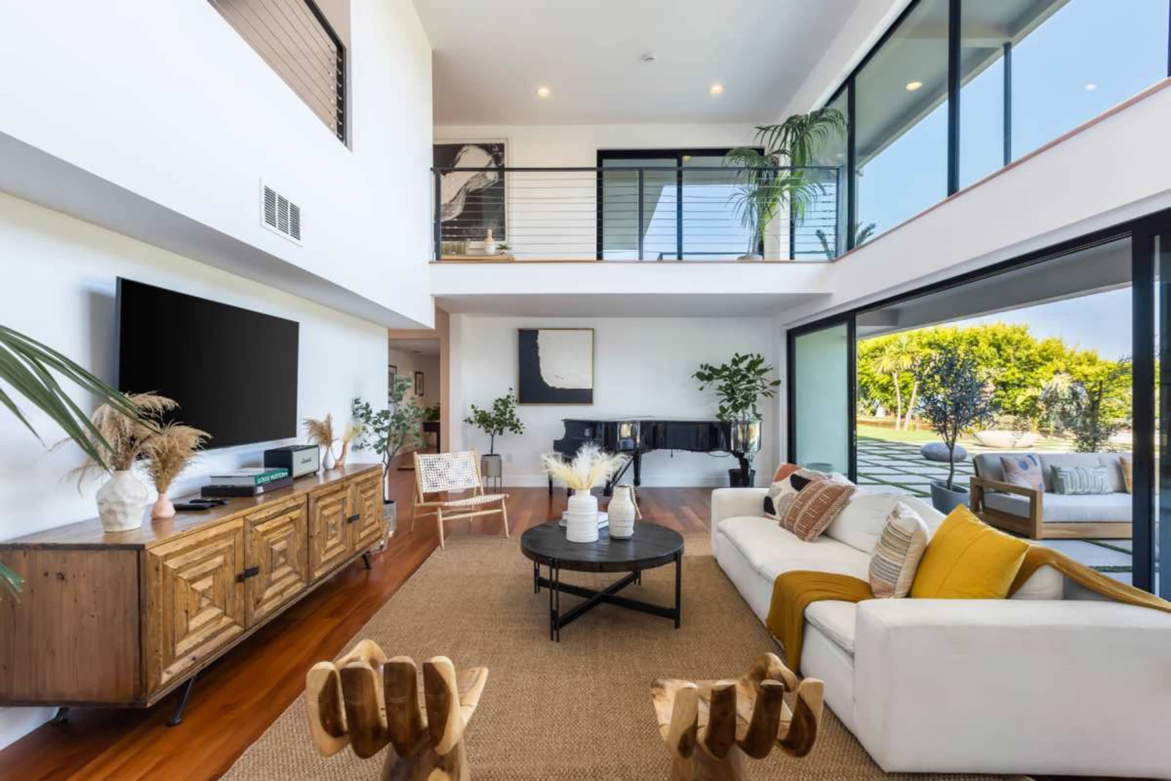 A modern living room with large windows, a wooden coffee table, a white sofa with yellow and beige pillows, and indoor plants.