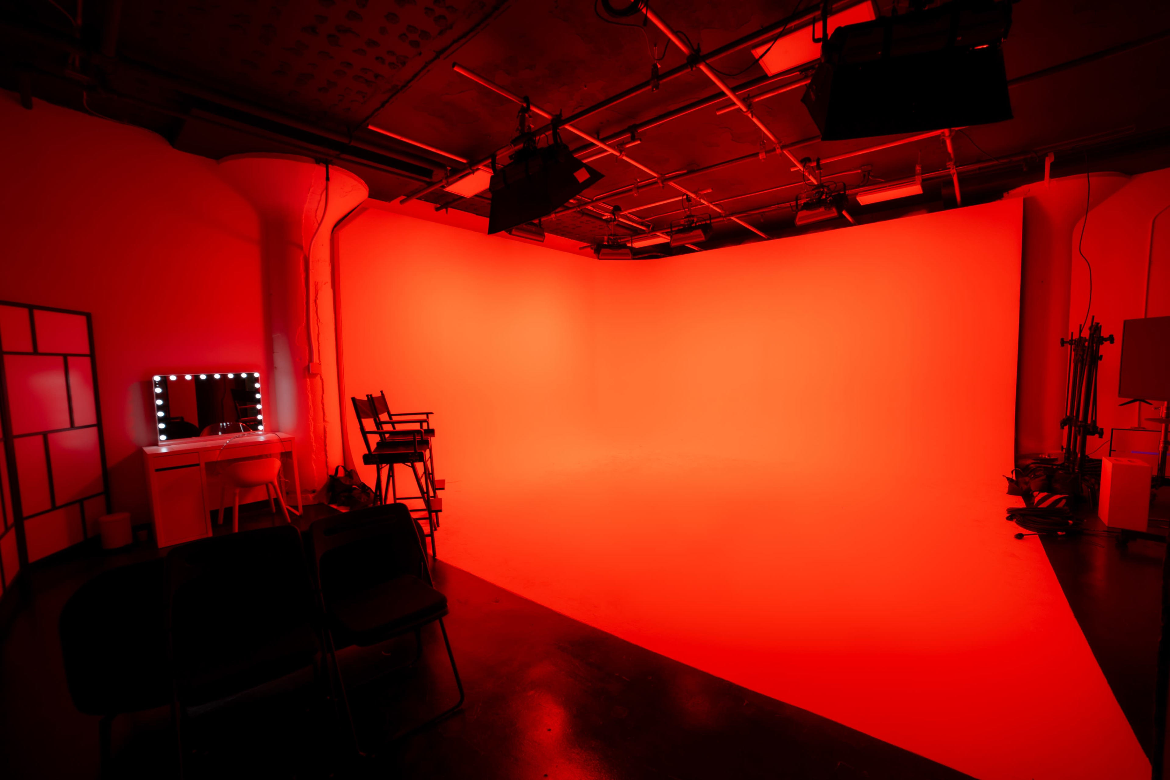 A photography studio with a solid red backdrop, a makeup vanity with lights, and a director's chair next to several black chairs.