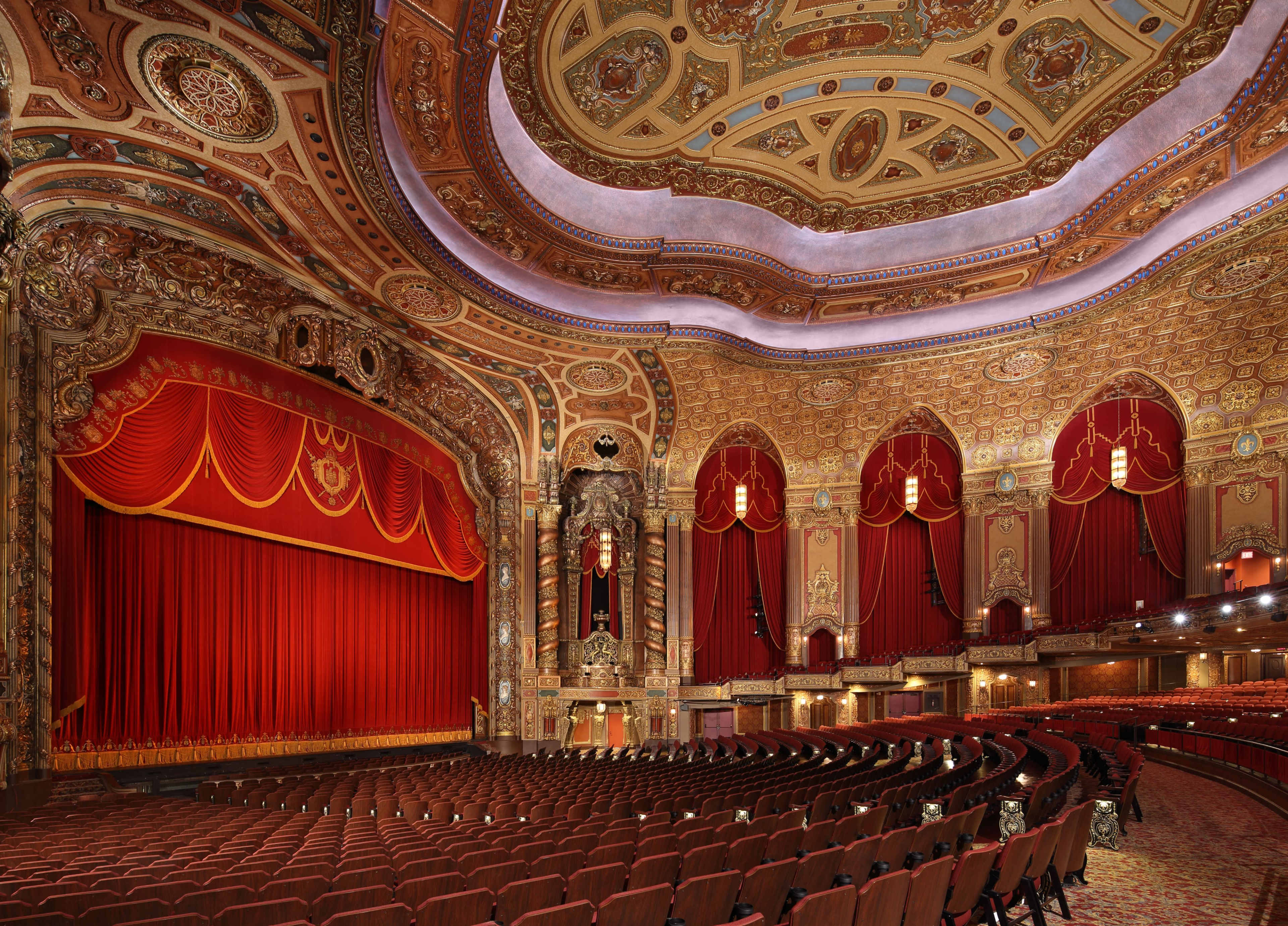The image shows an ornate theater interior with a red curtain, intricate ceiling designs, and rows of maroon seats.