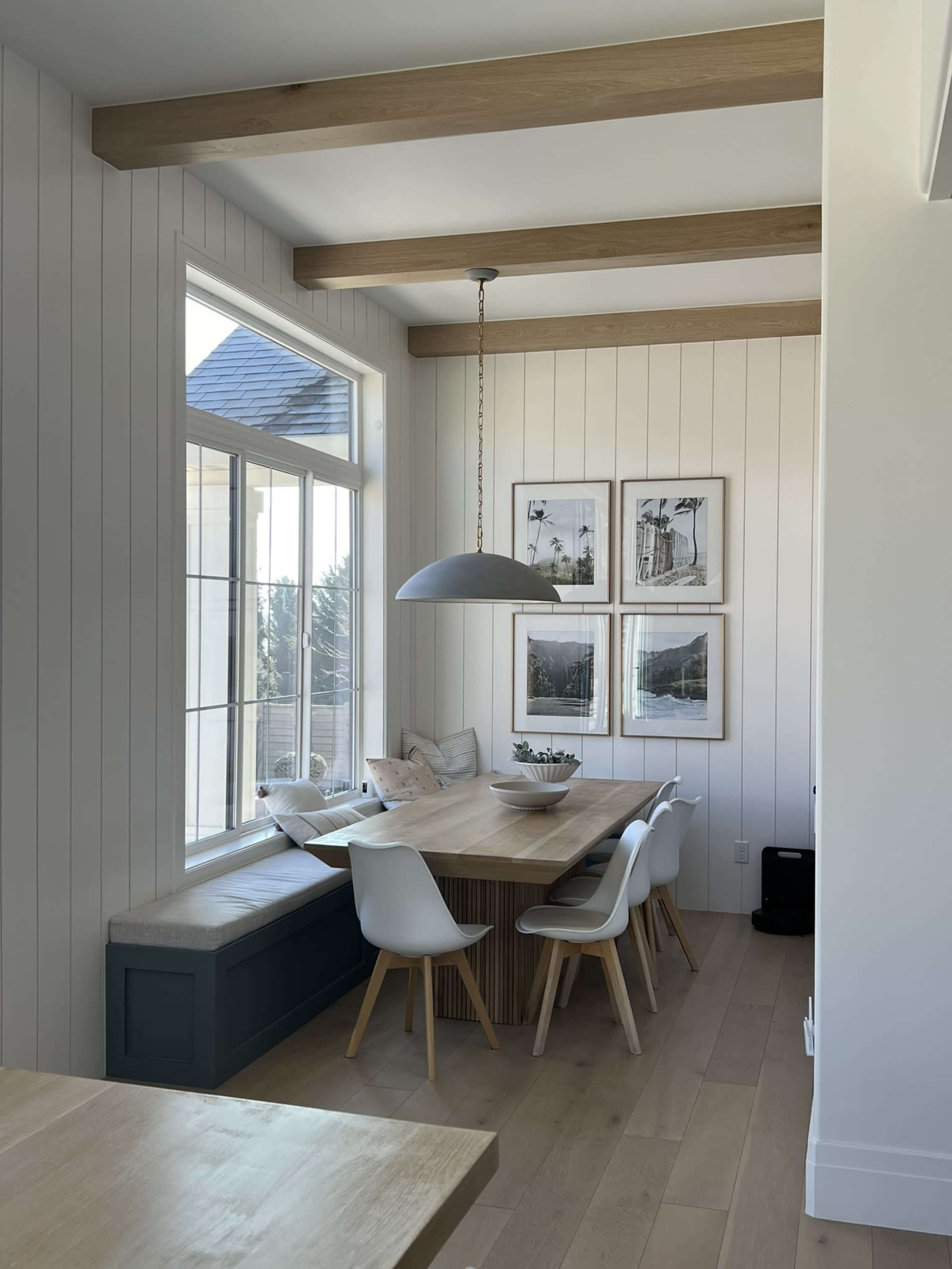 A dining area features a wooden table surrounded by white chairs, with a bench beneath a large window and framed photographs on the wall.