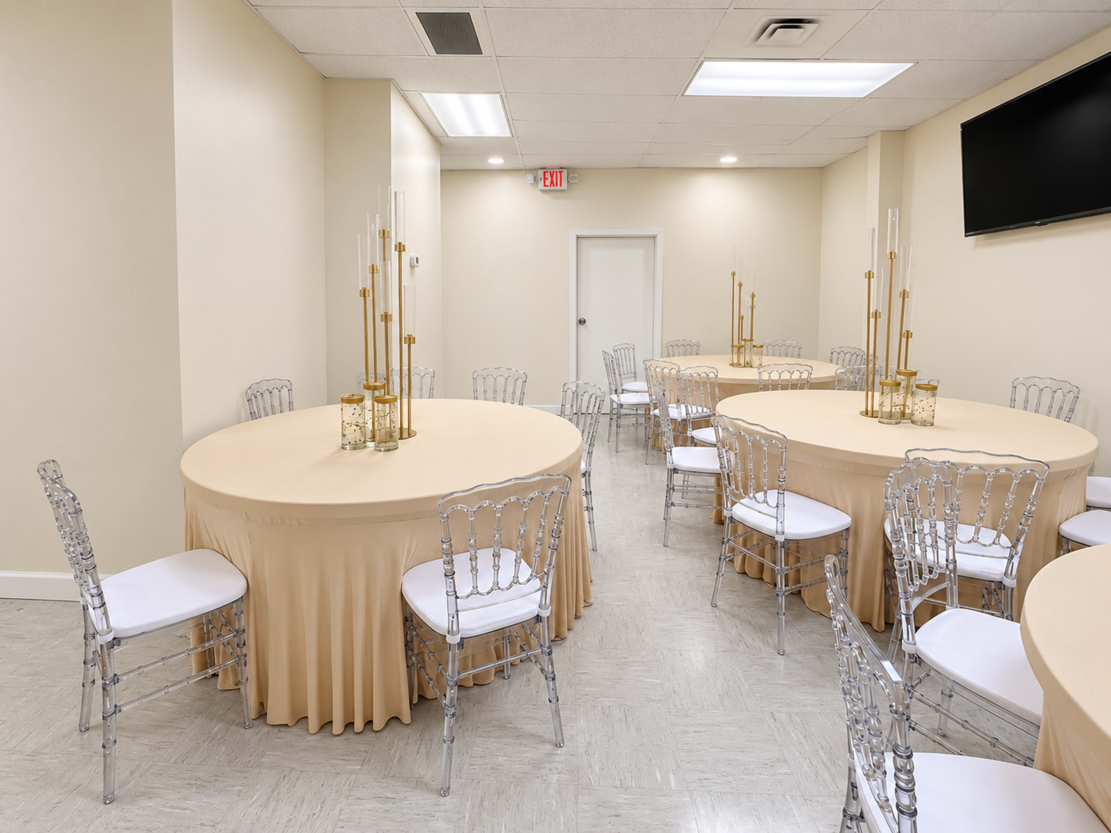 The image shows a well-lit event room with several round tables covered in beige tablecloths and surrounded by clear acrylic chairs.