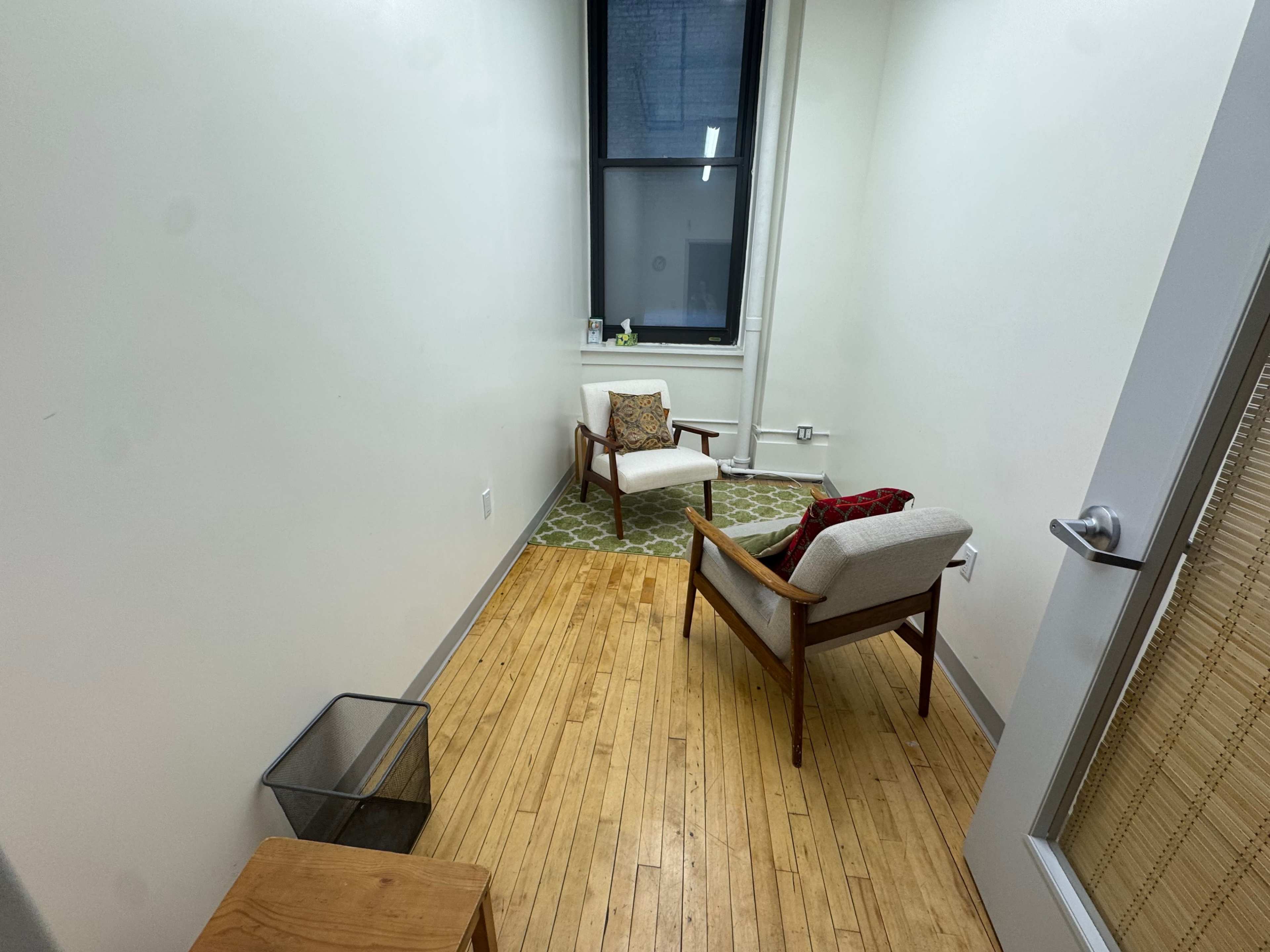 The image shows a small room with two armchairs, a rug on the floor, a wastebasket, and a window.