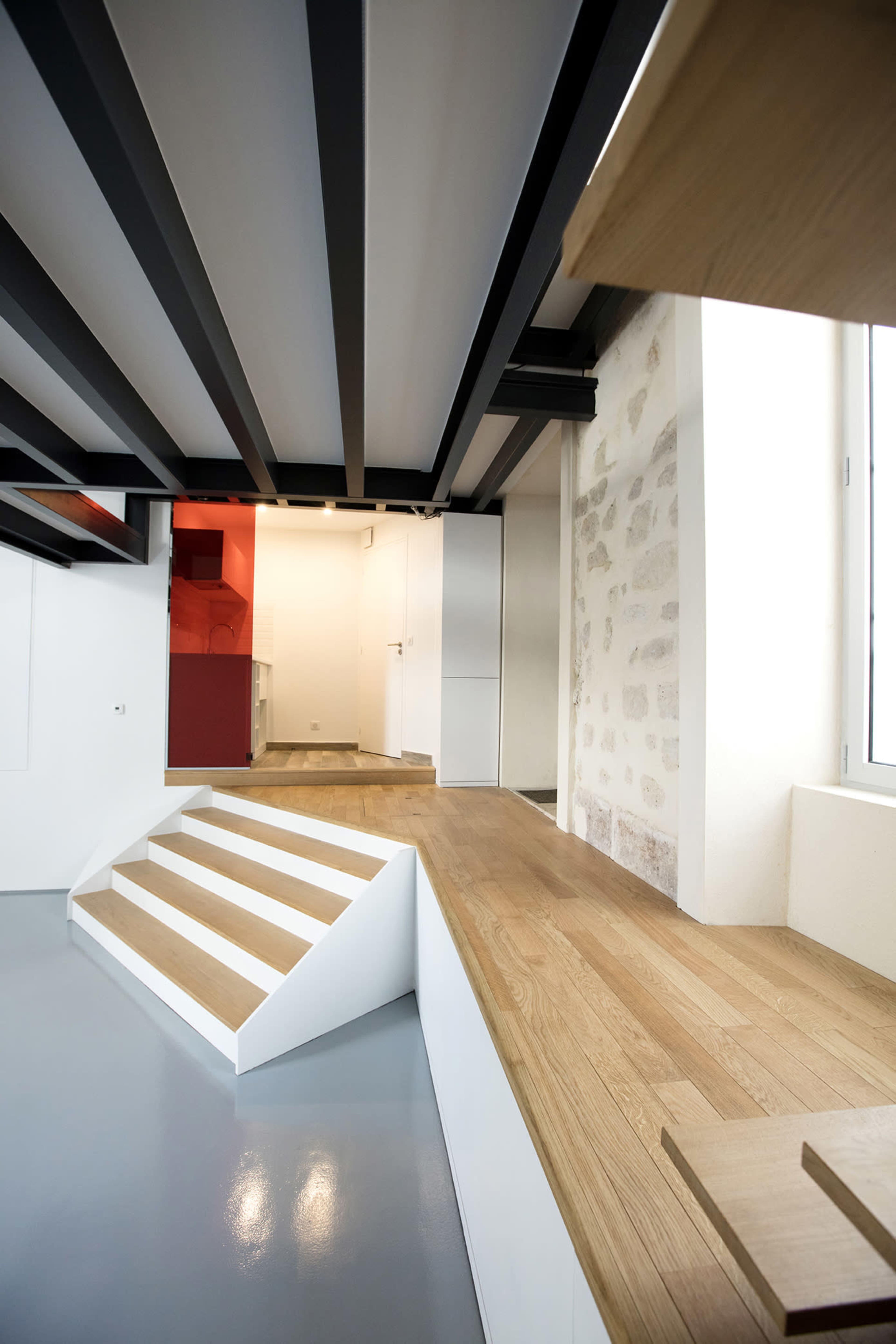 The image shows a modern interior space with a combination of wooden flooring, gray painted walls, and black exposed beams, featuring a set of white stairs leading to a raised platform.