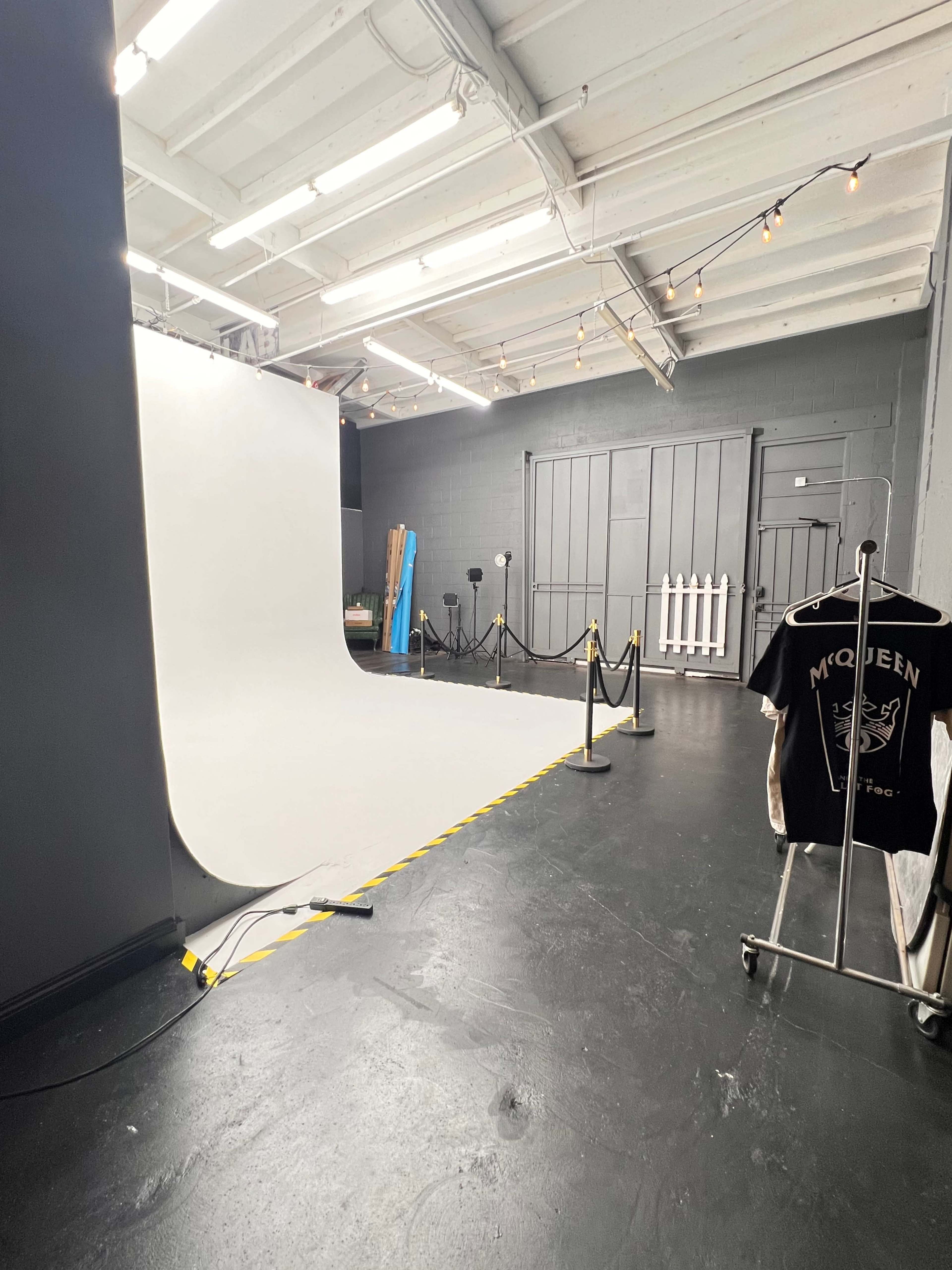 The image shows a photography studio with a white backdrop, black floor, and a clothing rack displaying a T-shirt, surrounded by black ropes and a wooden partition.