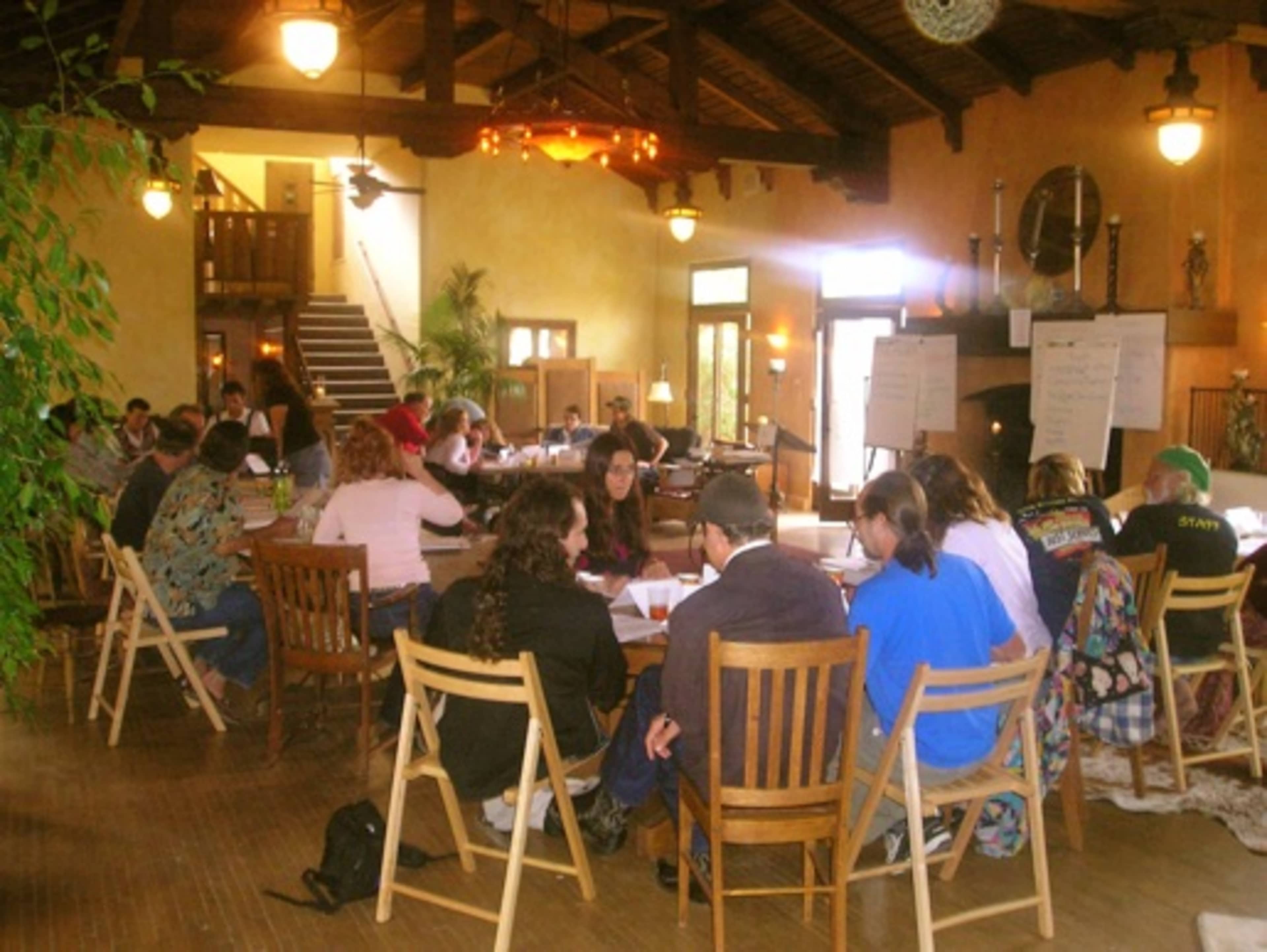 A group of people is seated around tables in a spacious room with high ceilings, participating in a meeting or workshop.