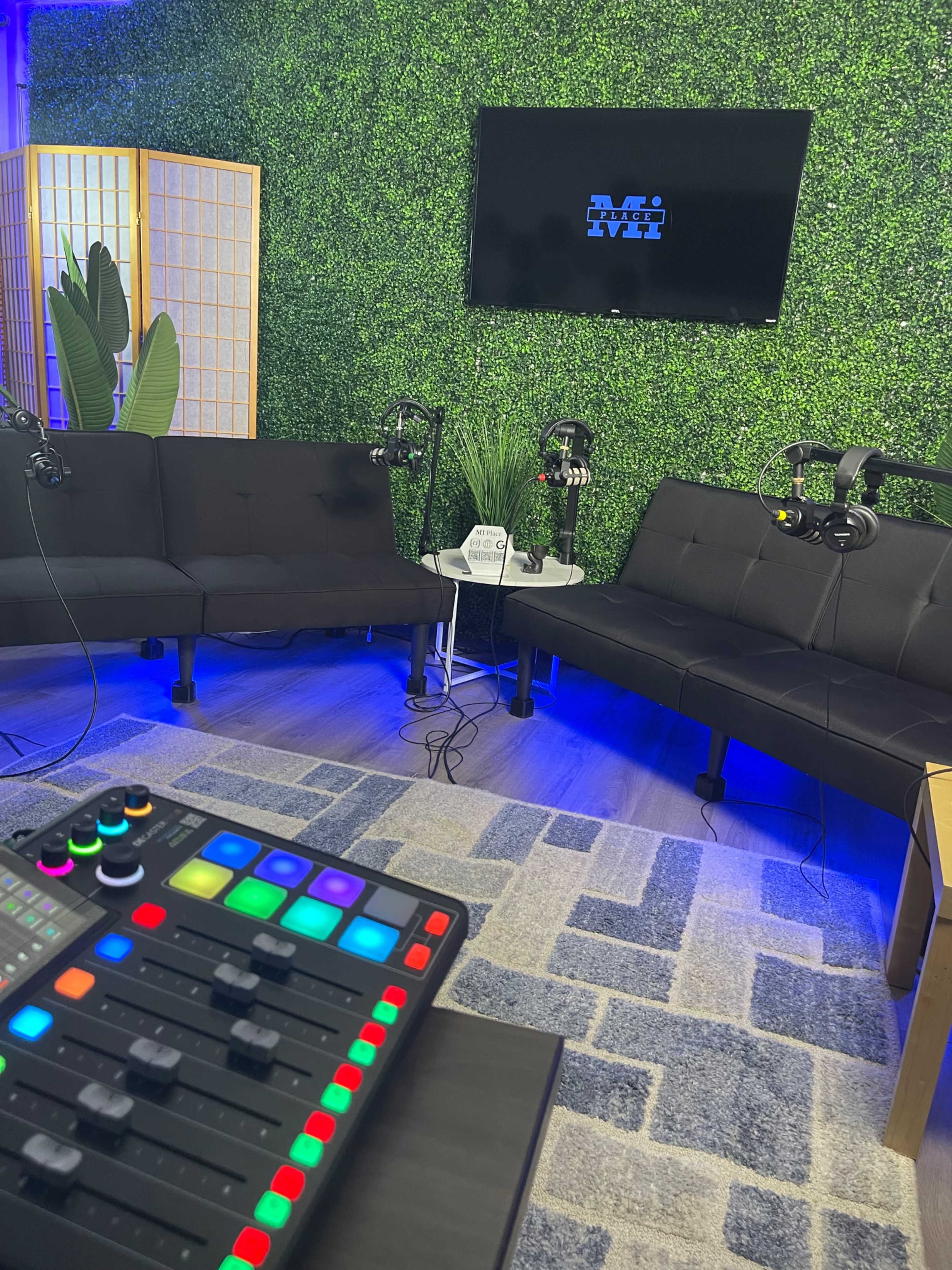 A recording studio setup with black couches, microphones on stands, a TV display on a green wall, and a colorful audio mixer in the foreground.
