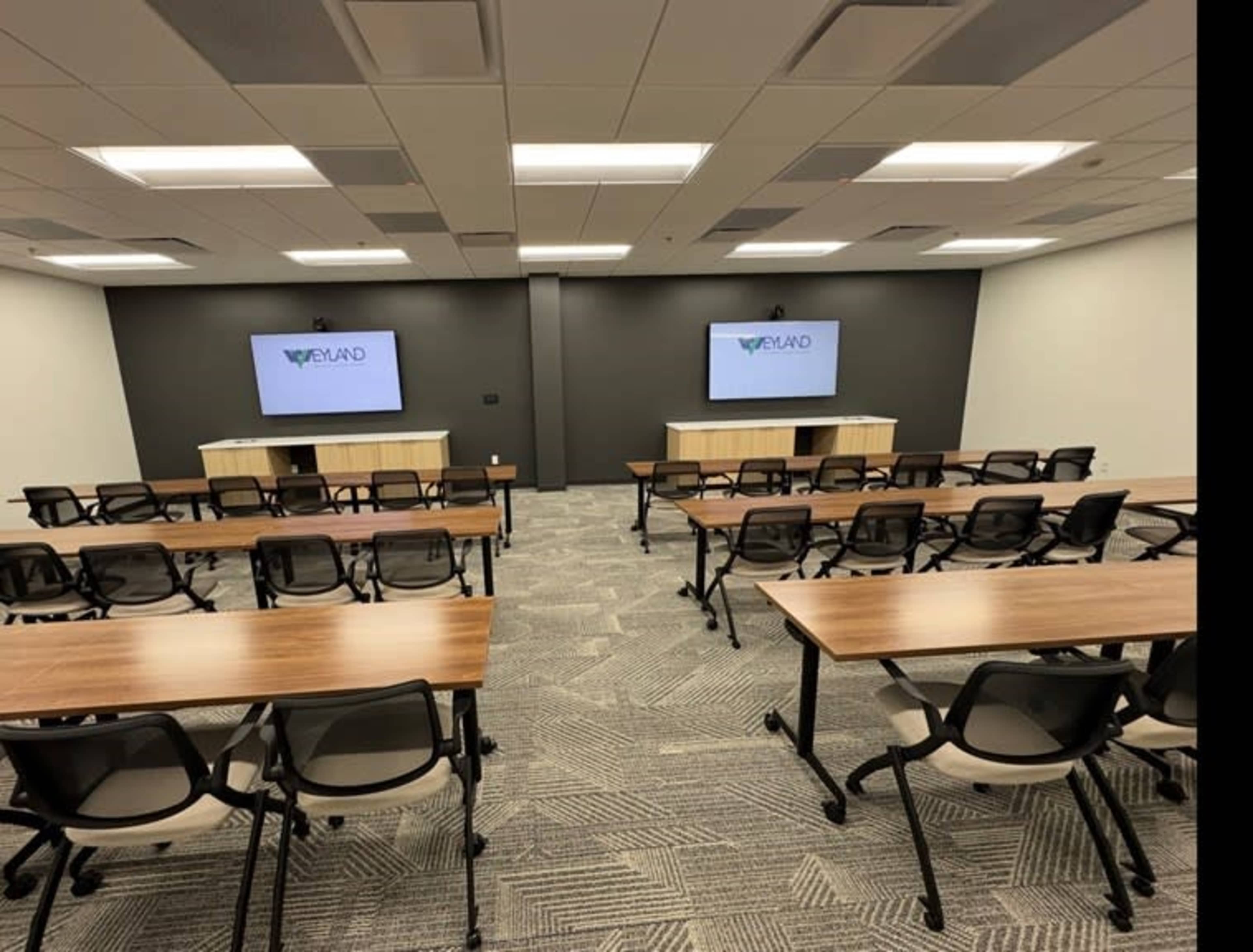 The image shows a conference room equipped with two screens, rows of tables, and chairs arranged for a meeting.
