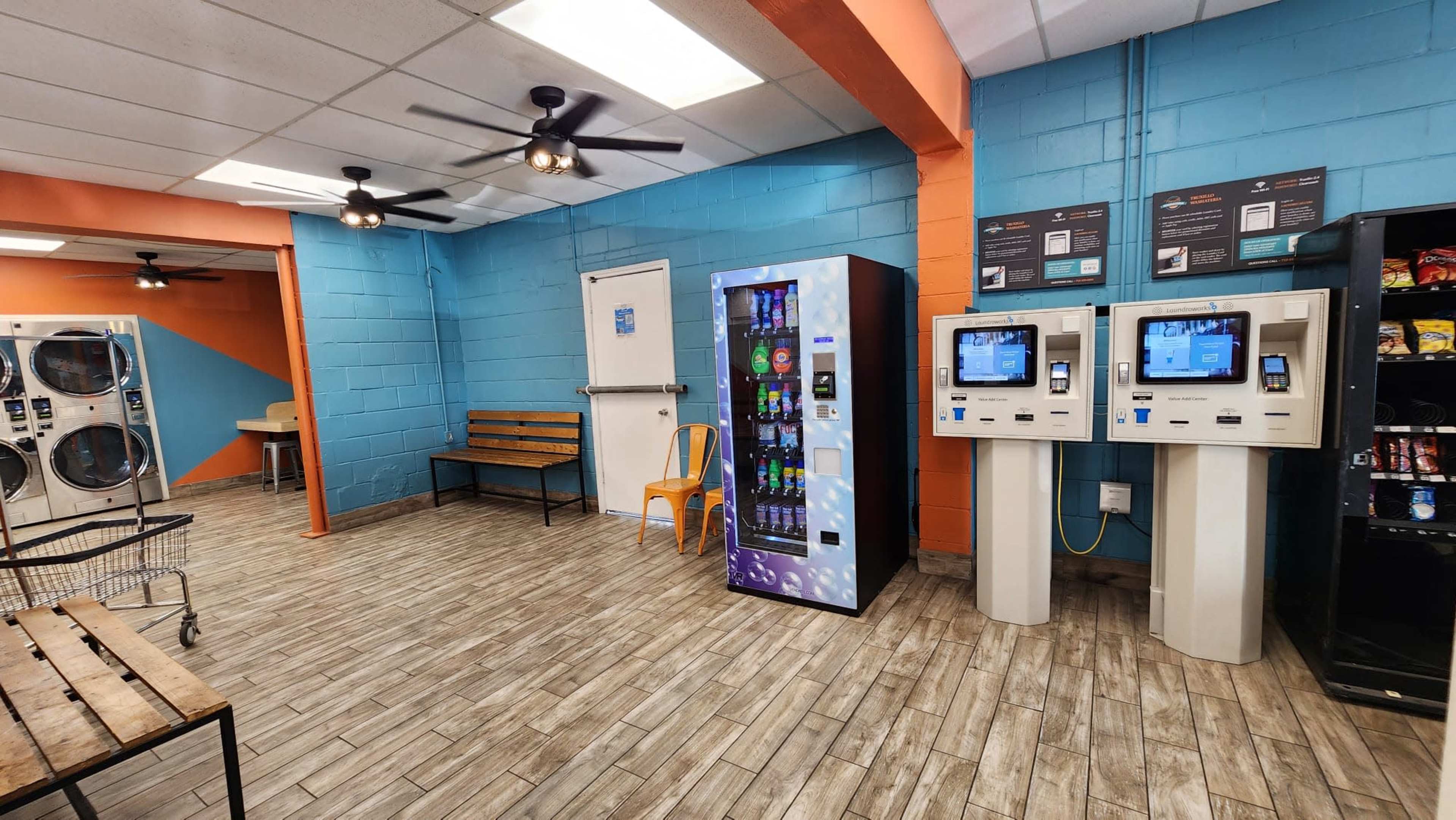 A laundromat interior featuring vending machines and self-service kiosks against a backdrop of blue and orange walls.