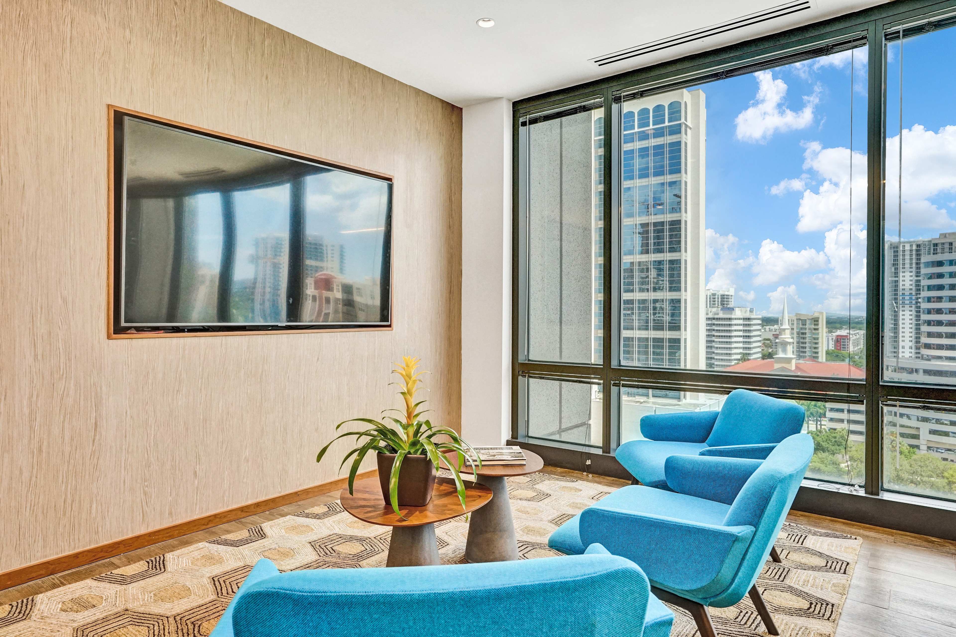 A modern lounge area features two blue chairs, a small table with a potted plant, and a large window overlooking a city skyline.