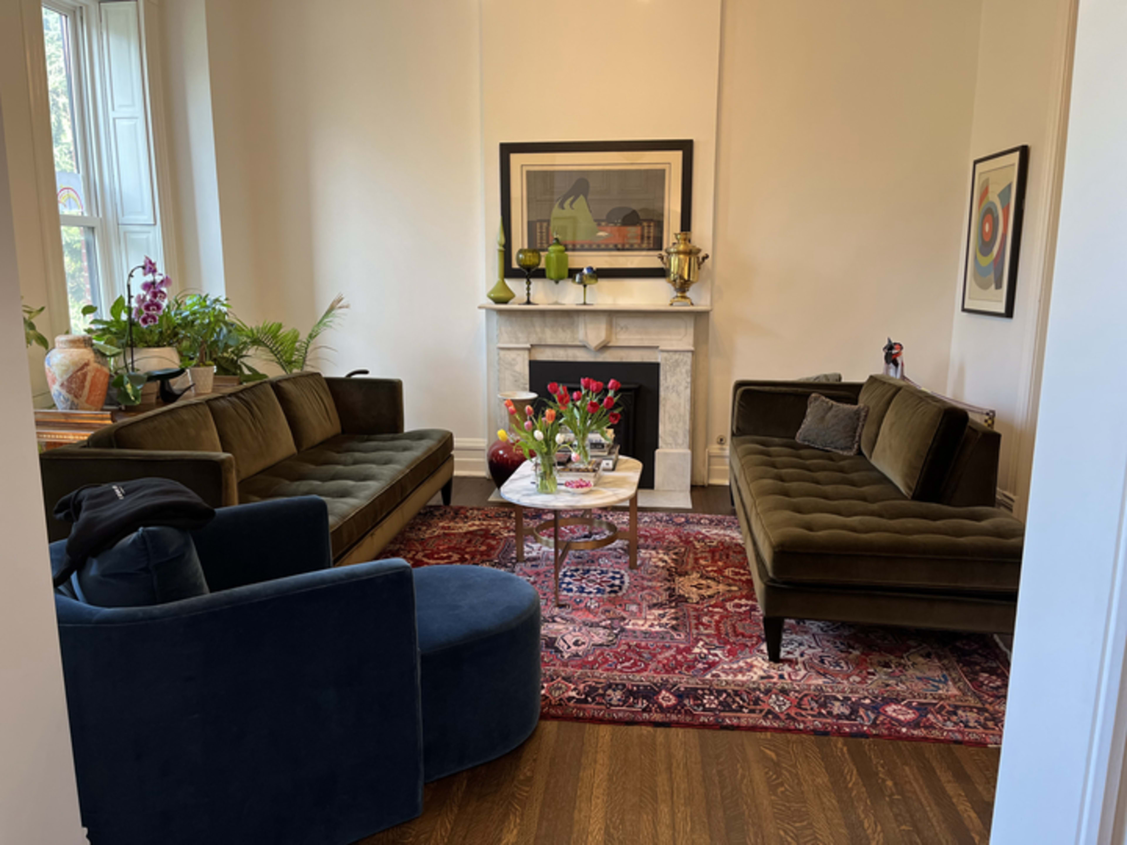 A living room with two green sofas, a round coffee table on a patterned rug, and a fireplace adorned with plants and decorative items.