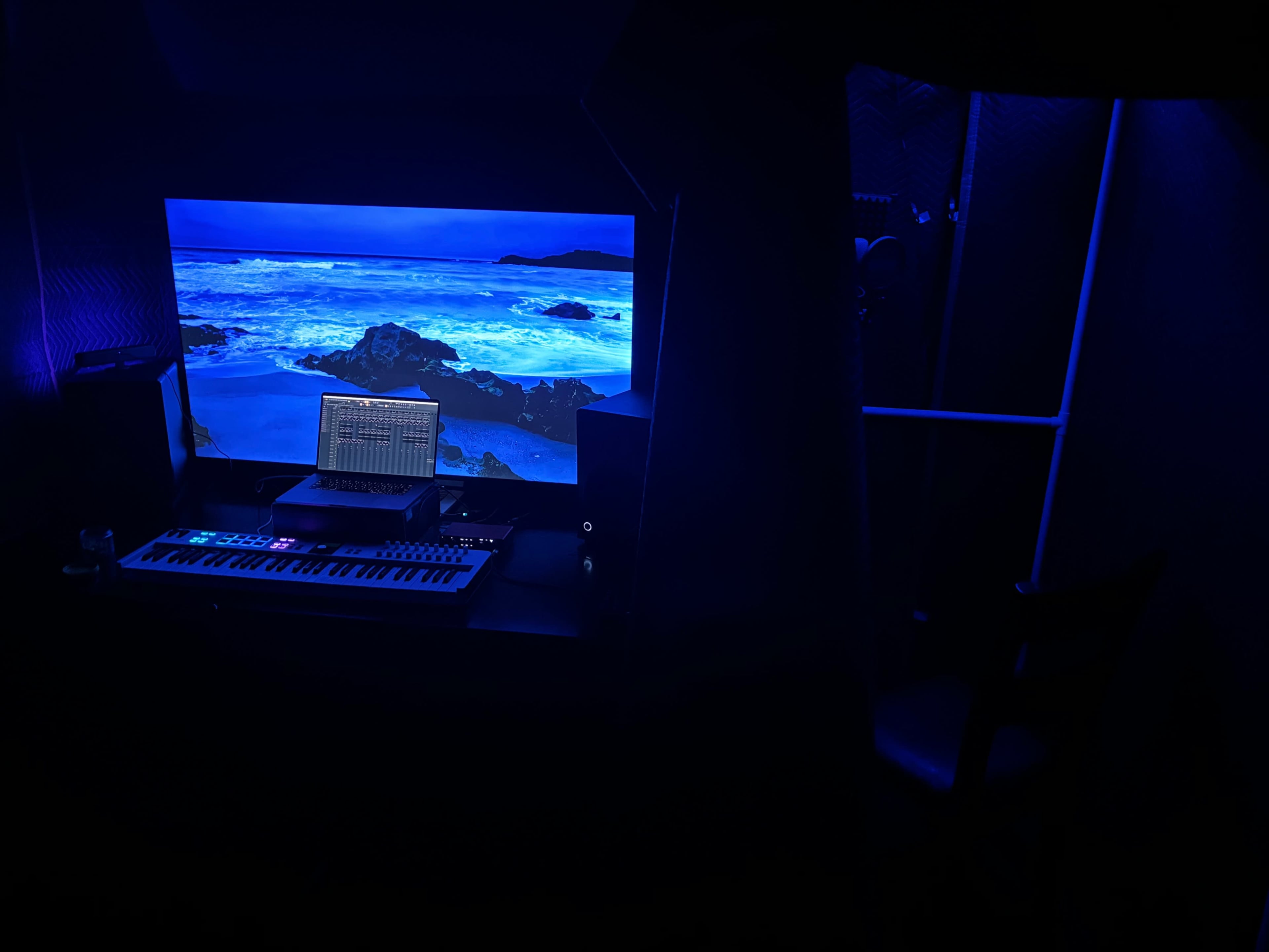 A dimly lit music production setup featuring a keyboard, a laptop displaying audio software, and a large screen with a coastal landscape.
