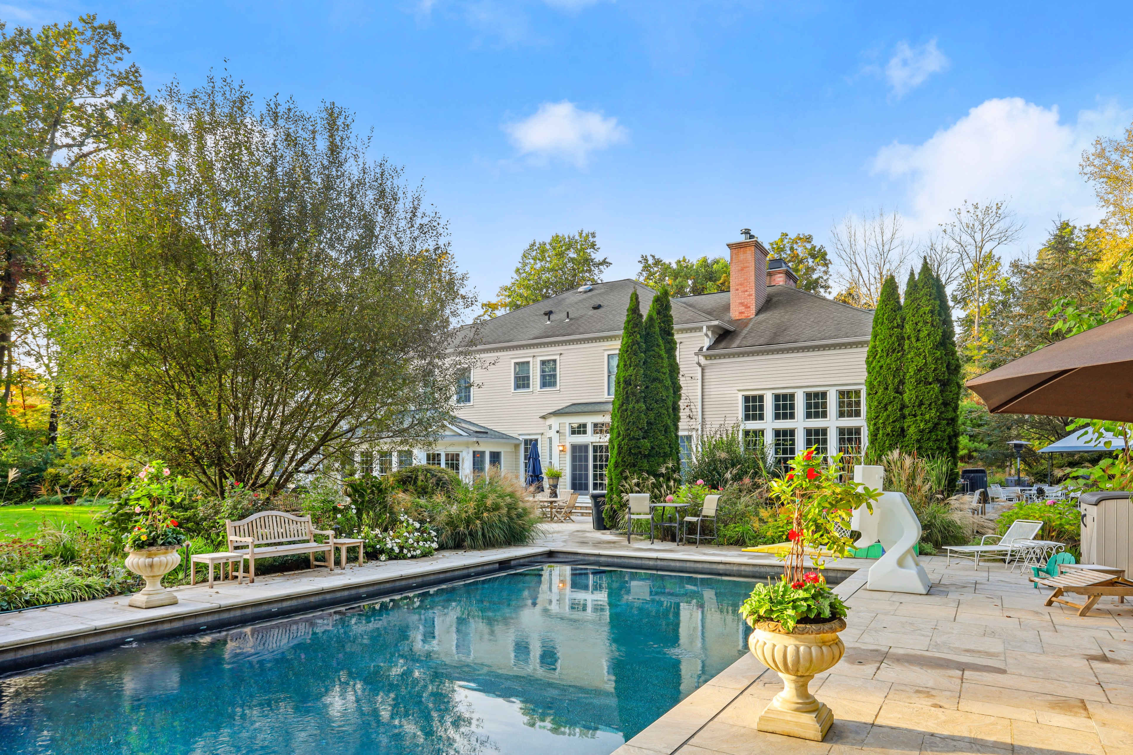 The image shows a backyard with a swimming pool in the foreground, a large house in the background, and landscaped gardens with trees and seating areas.