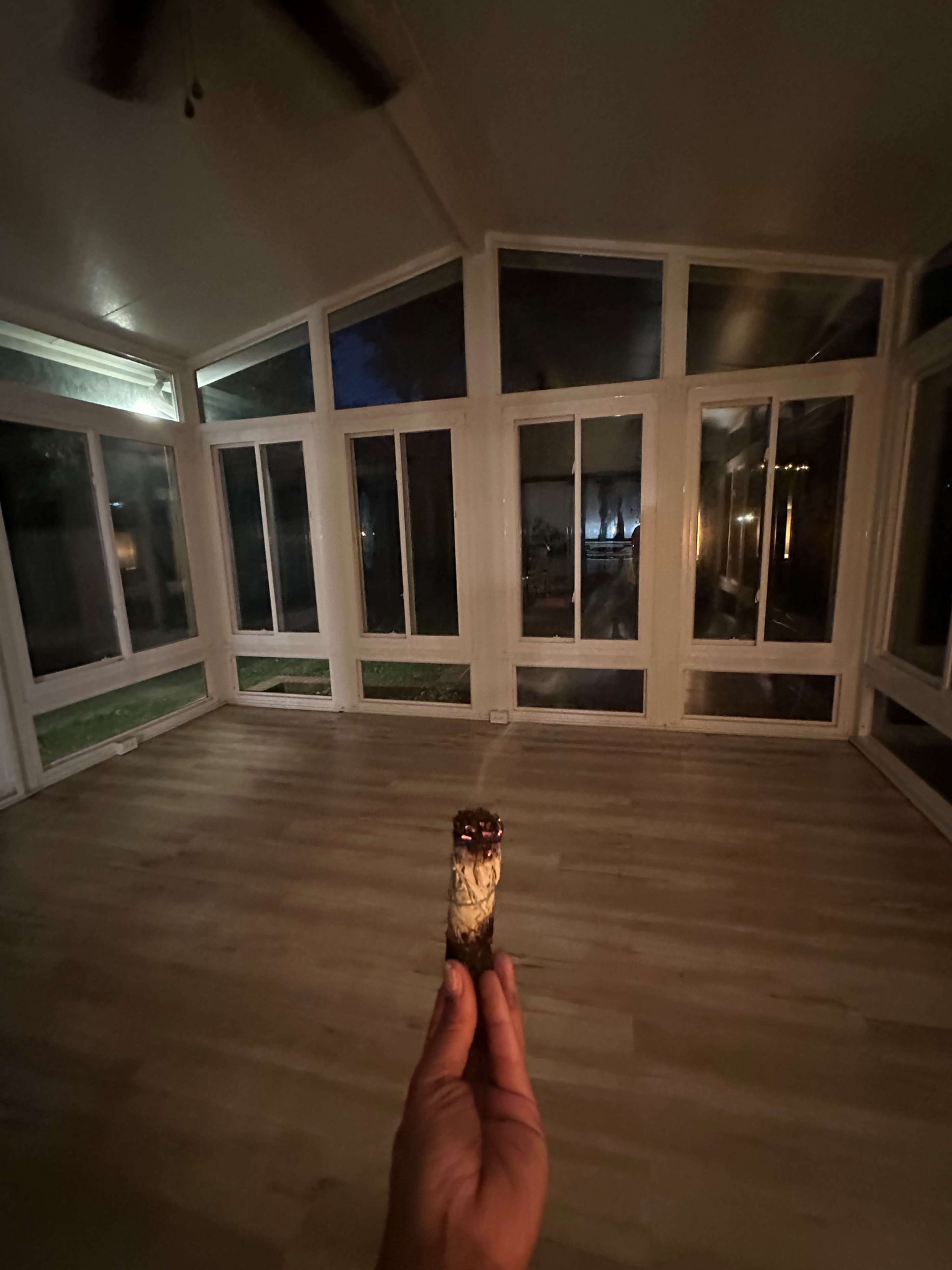 A person holds a lit cigar in an empty sunroom with large windows and wooden flooring.